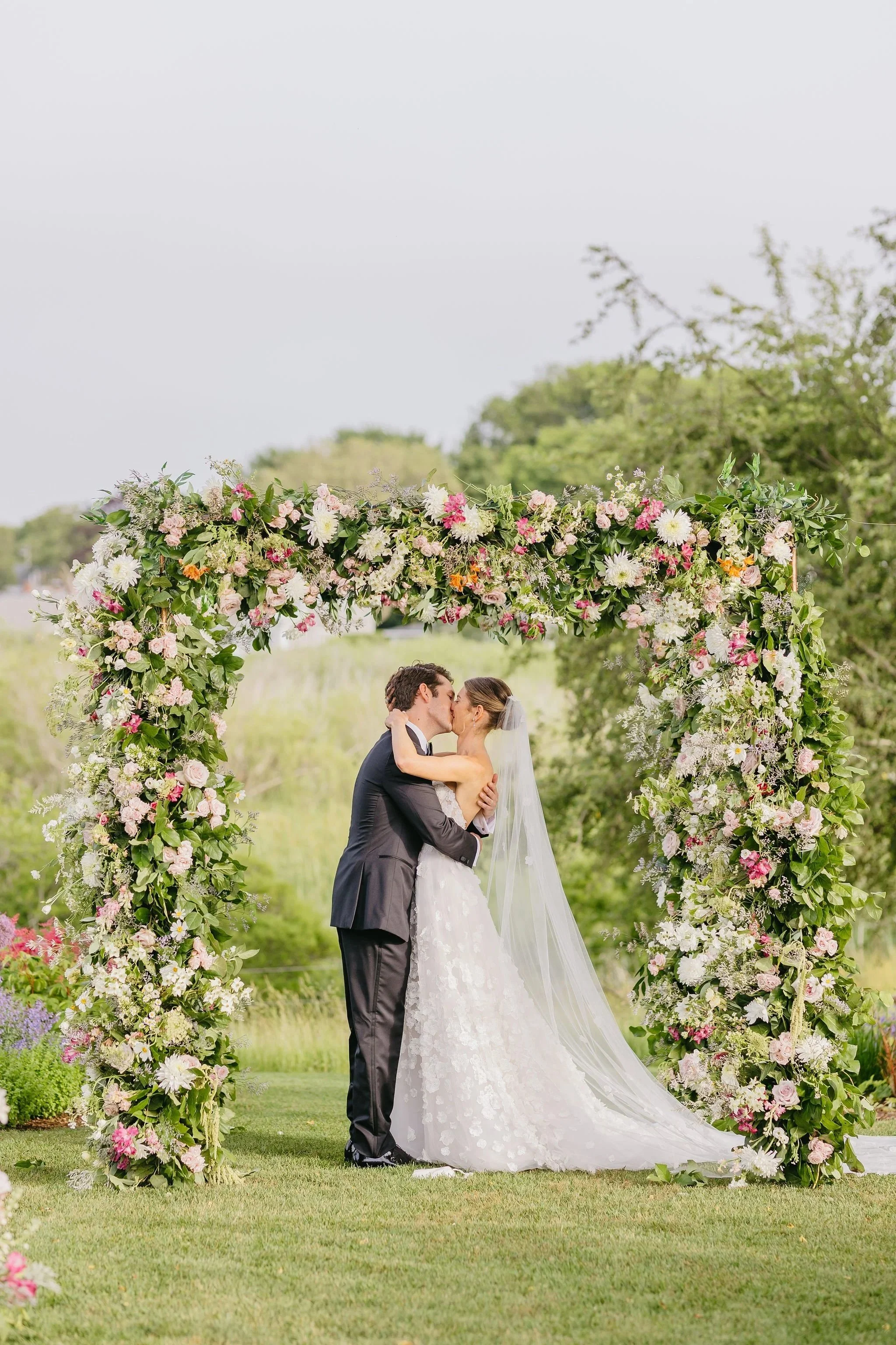 Nantucket wedding garden ceremony. A bride and groom kissing under a floral wedding arch outdoors with green trees and grass.