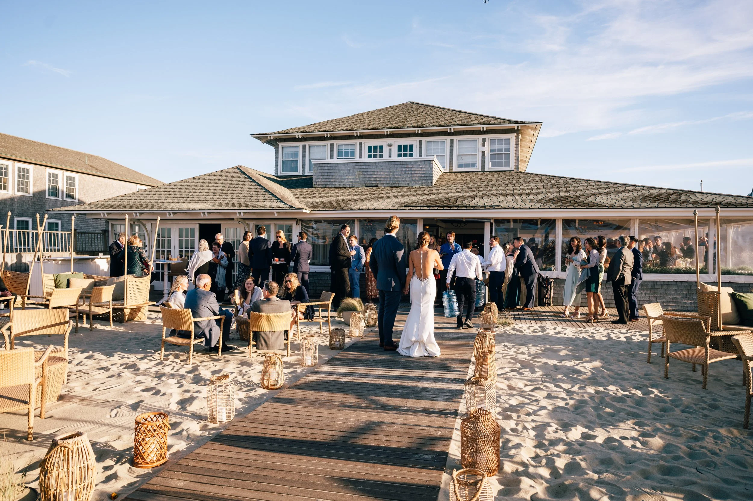 Galley Beach wedding reception. People gathered on a beach for a wedding reception near a large beach house, with some sitting on chairs, others standing and talking, decorated with lanterns and candles, under clear skies.