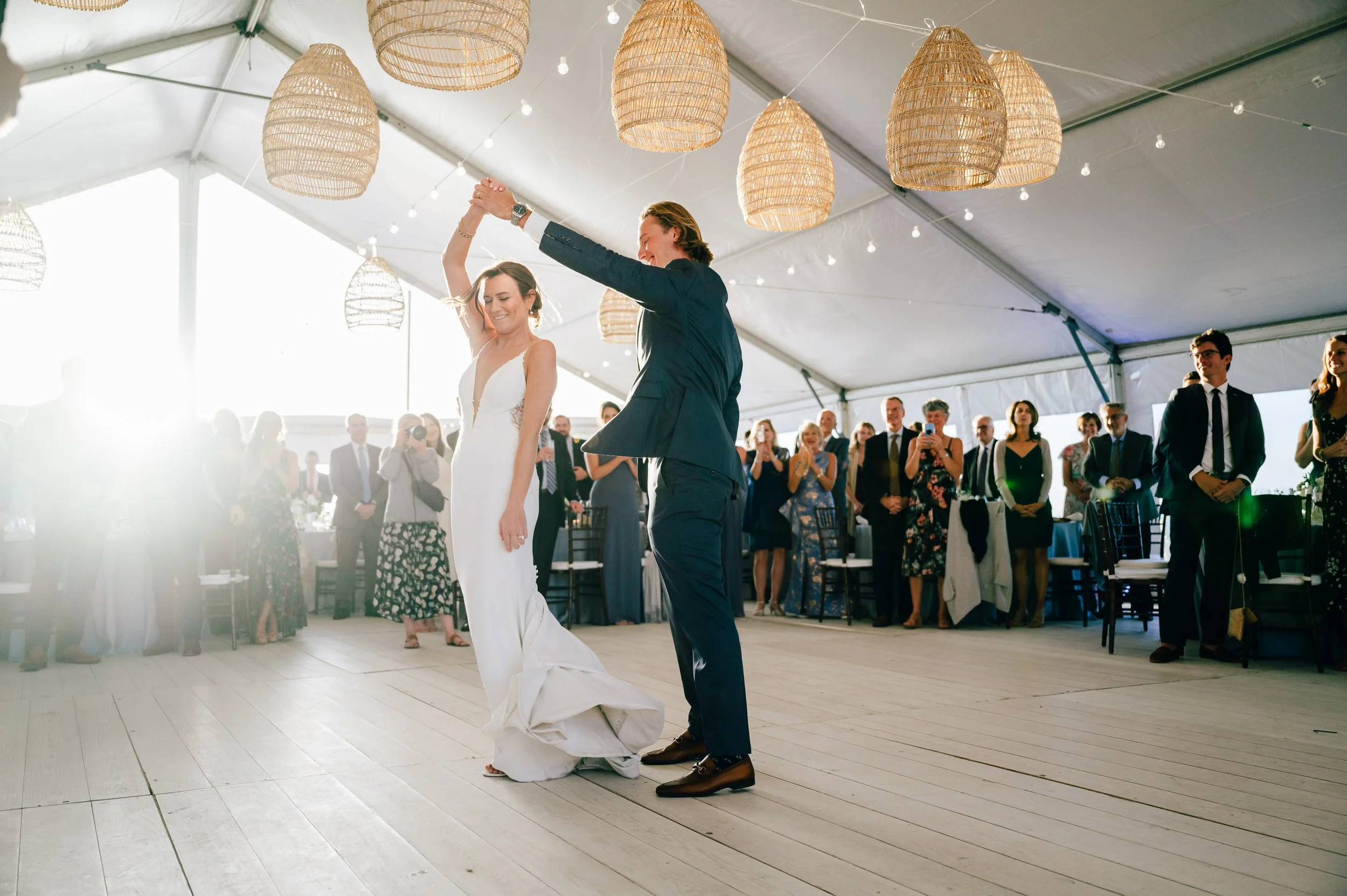 Galley Beach Nantucket Wedding. A bride and groom are dancing during their wedding reception under a tent with hanging wicker lamps. Guests are watching and smiling in the background.