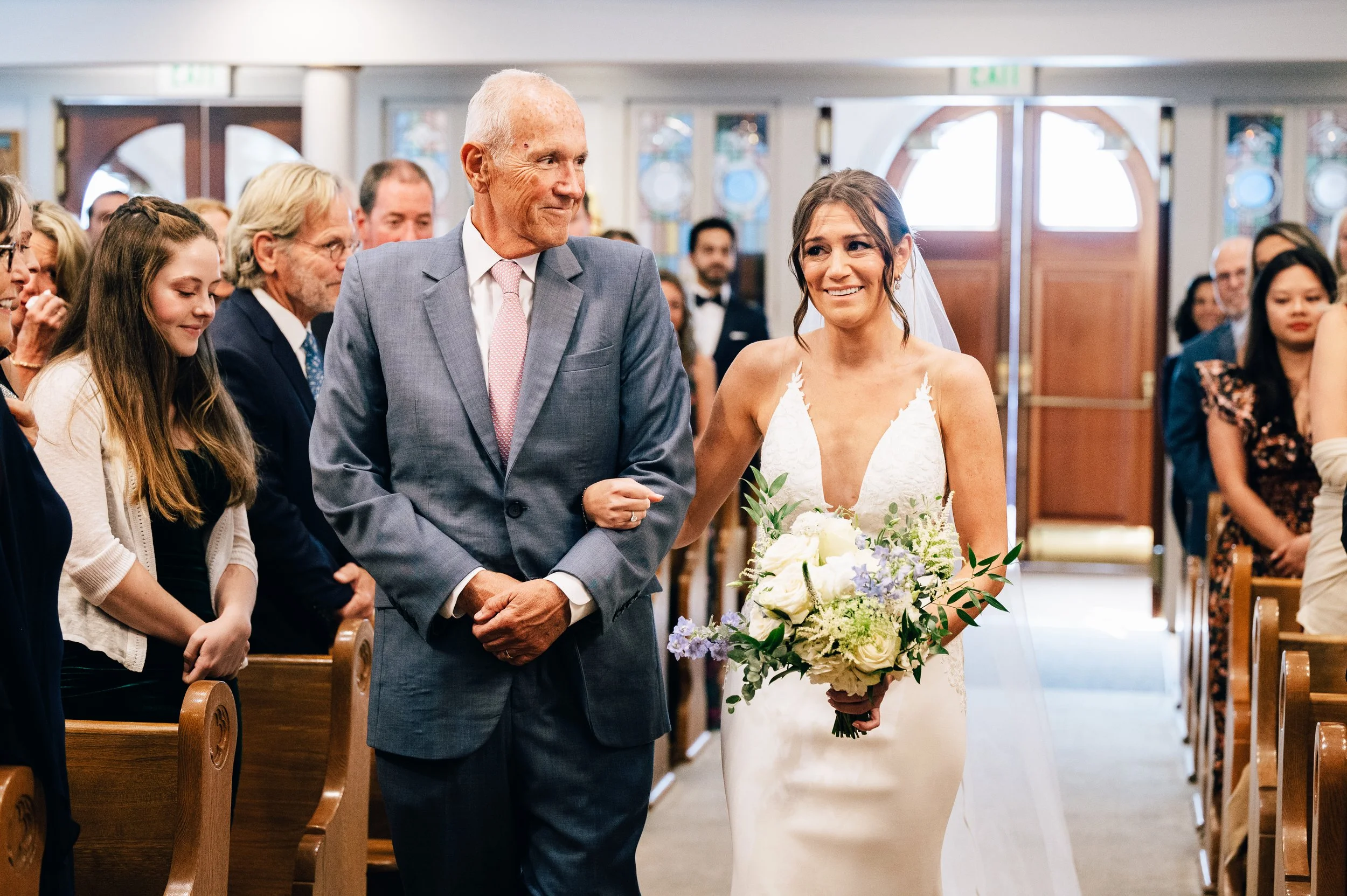 St Marys Nantucket Wedding ceremony. A bride in a white wedding dress holding a bouquet of white and purple flowers, walking down the aisle with an older man in a gray suit, during a wedding ceremony in a church filled with guests.