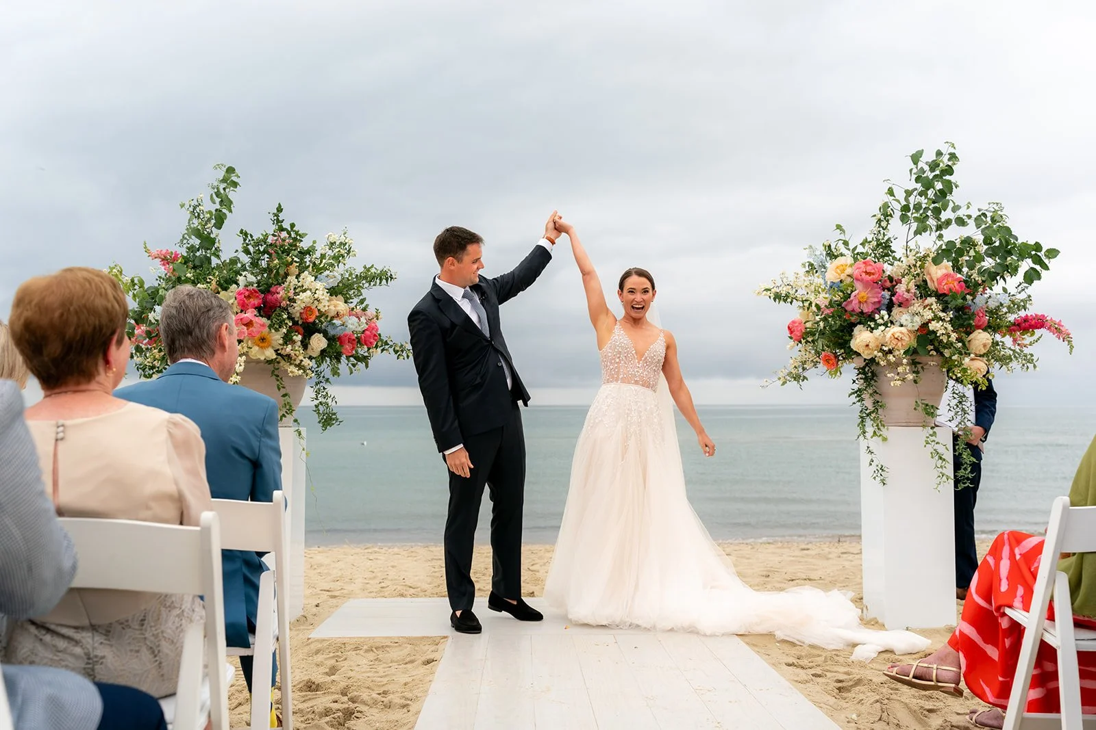A newlywed couple holding hands and celebrating their wedding ceremony on a beach. The bride is wearing a white wedding gown, and the groom is dressed in a dark suit. Guests are seated on white chairs, and large floral arrangements decorate the ceremony area against a backdrop of the ocean and cloudy sky.