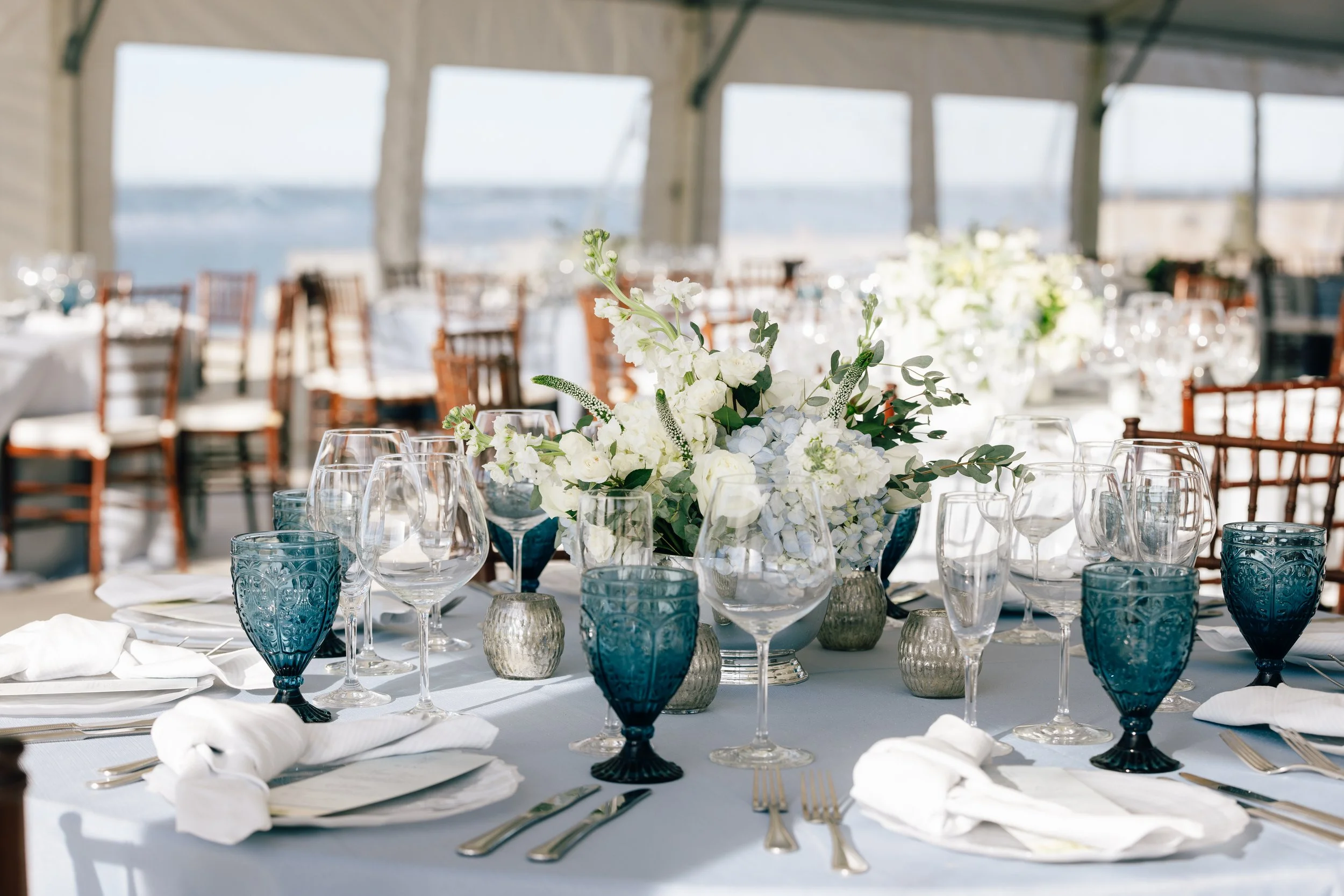 Elegant banquet table set with white flowers in a centerpiece, surrounded by blue and clear glasses, white napkins, and silverware, under a tent with a scenic ocean view in the background.