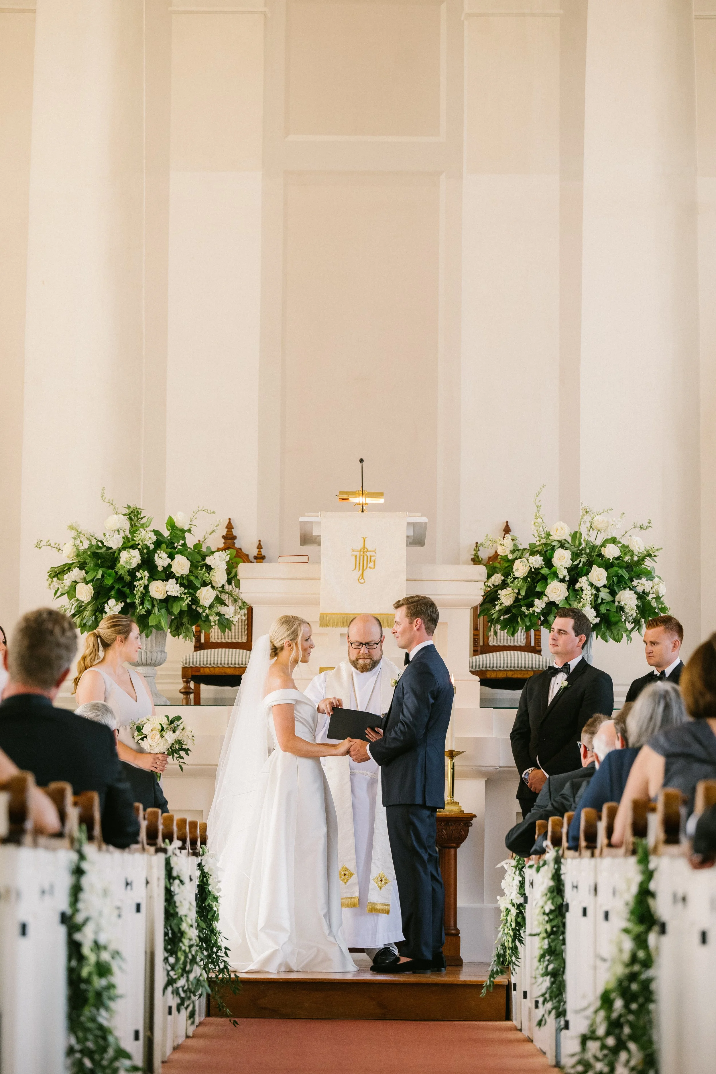First Congregational Church Nantucket Wedding. A bride and groom standing at the altar in a church during their wedding ceremony, surrounded by wedding party members and guests, with floral arrangements on either side of the aisle.
