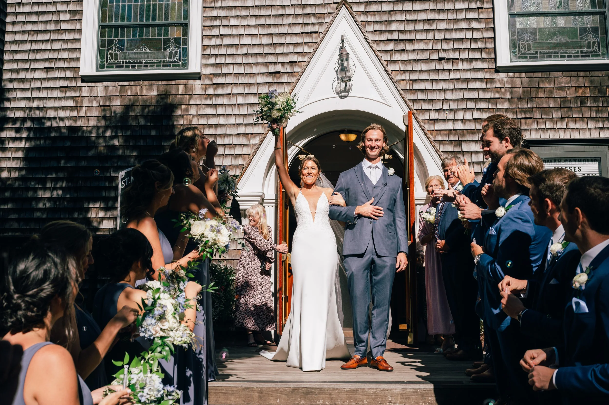 St Marys Nantucket Wedding Ceremony. Bride and groom exiting a church after wedding ceremony, surrounded by friends and family, with the bride holding a bouquet and wearing a white dress, and the groom in a gray suit.