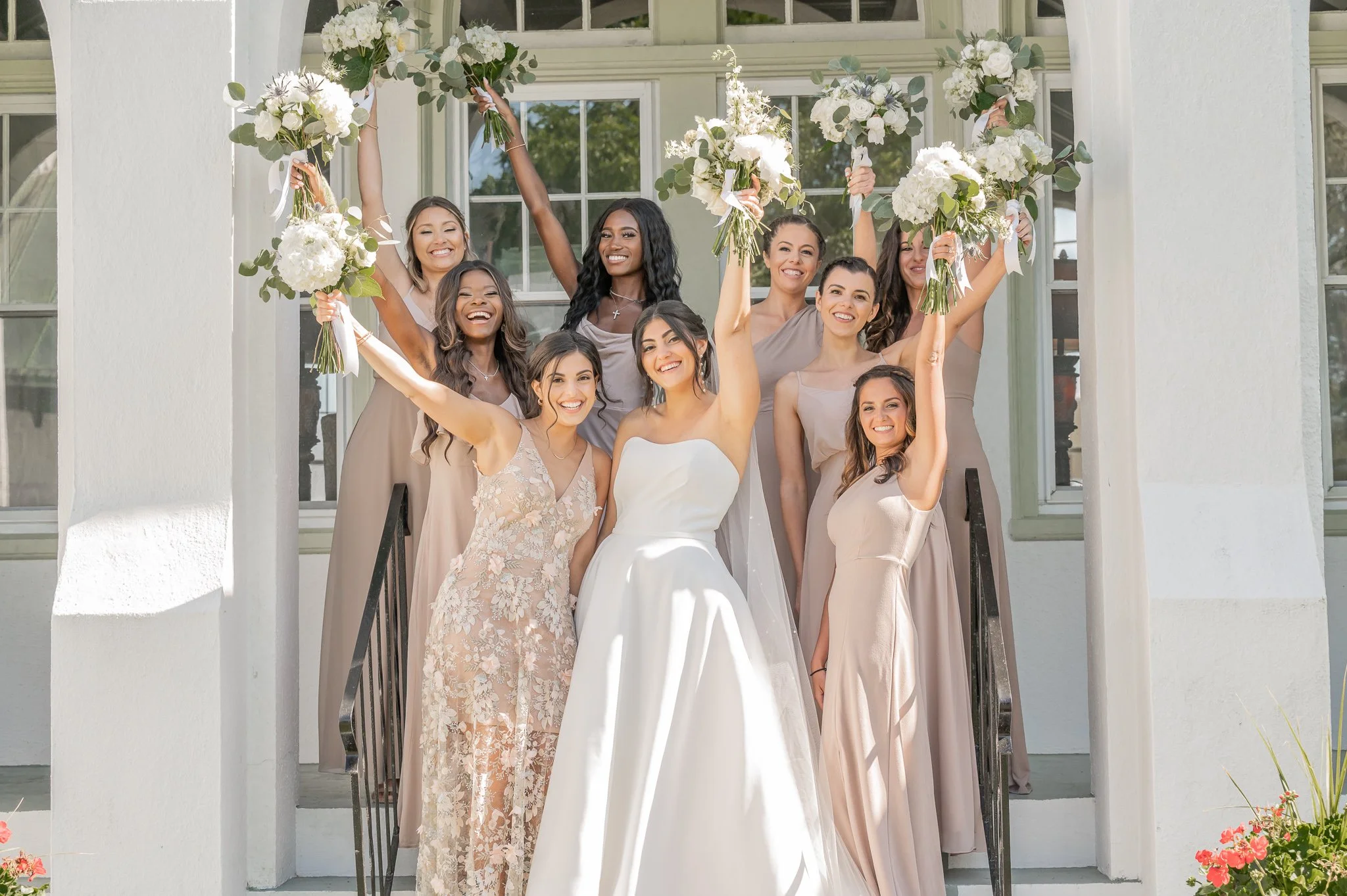 A group of women in beige dresses celebrating on stairs with bouquets of white flowers