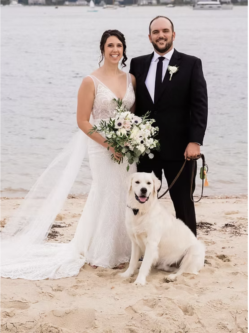 Brant point wedding. A bride and groom standing on a beach, with a golden retriever dog sitting in front of them. The bride is holding a bouquet of white and purple flowers, and they are smiling.
