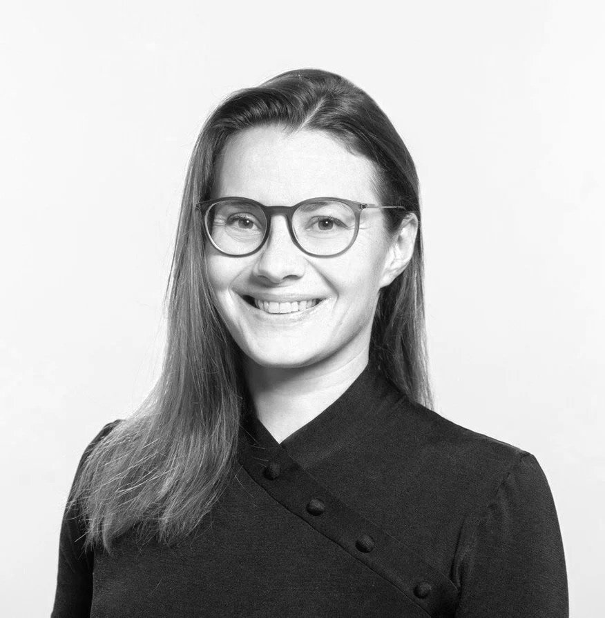 Black and white portrait of a woman with long hair, wearing glasses and a dark buttoned top, smiling at the camera against a plain background.