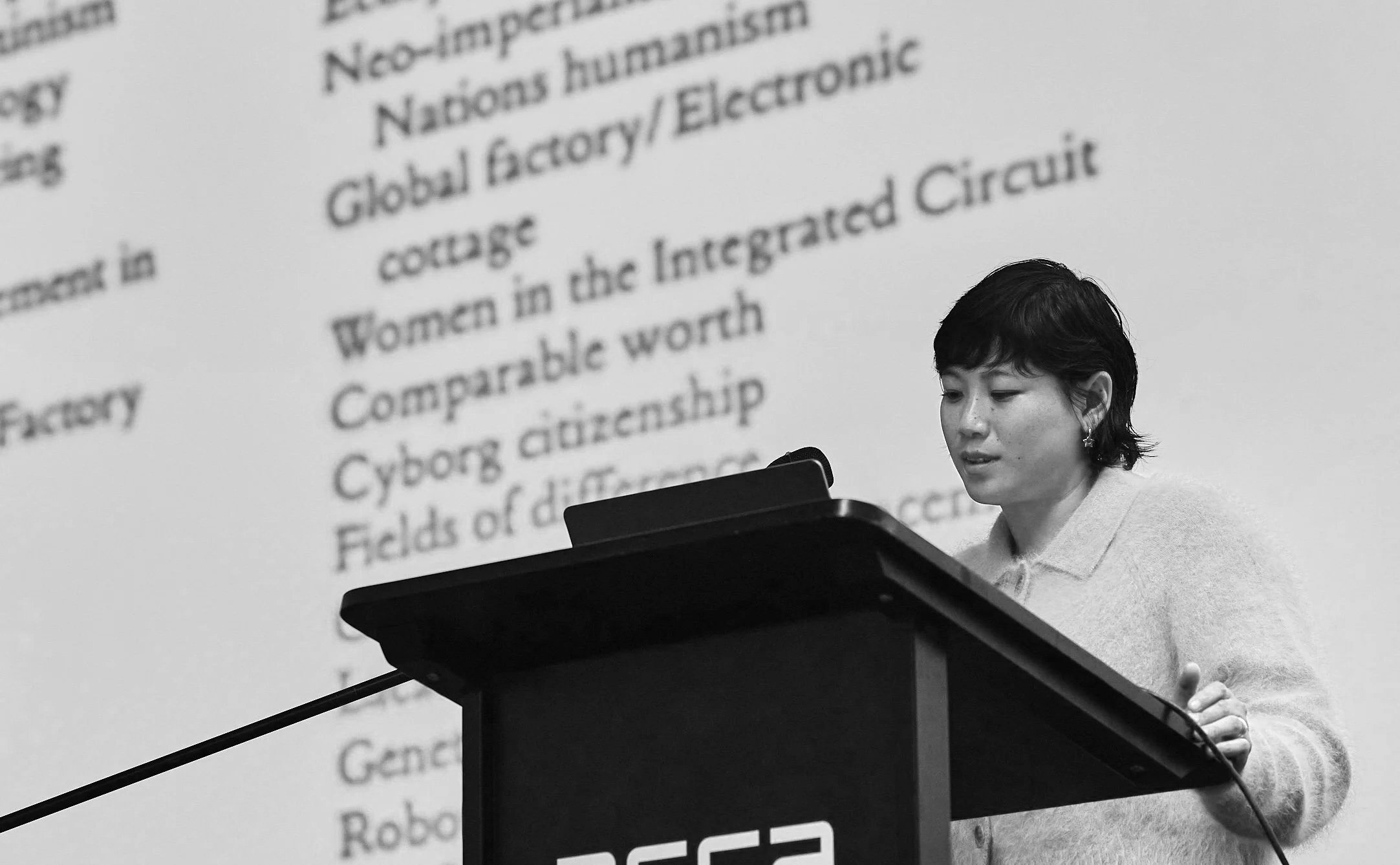 A woman with short dark hair and earrings reading or presenting from a document at a lectern, with a large, blurry projection screen behind her displaying text about nonimpairment, women in the integrated circuit habit, and cyber citizenship.