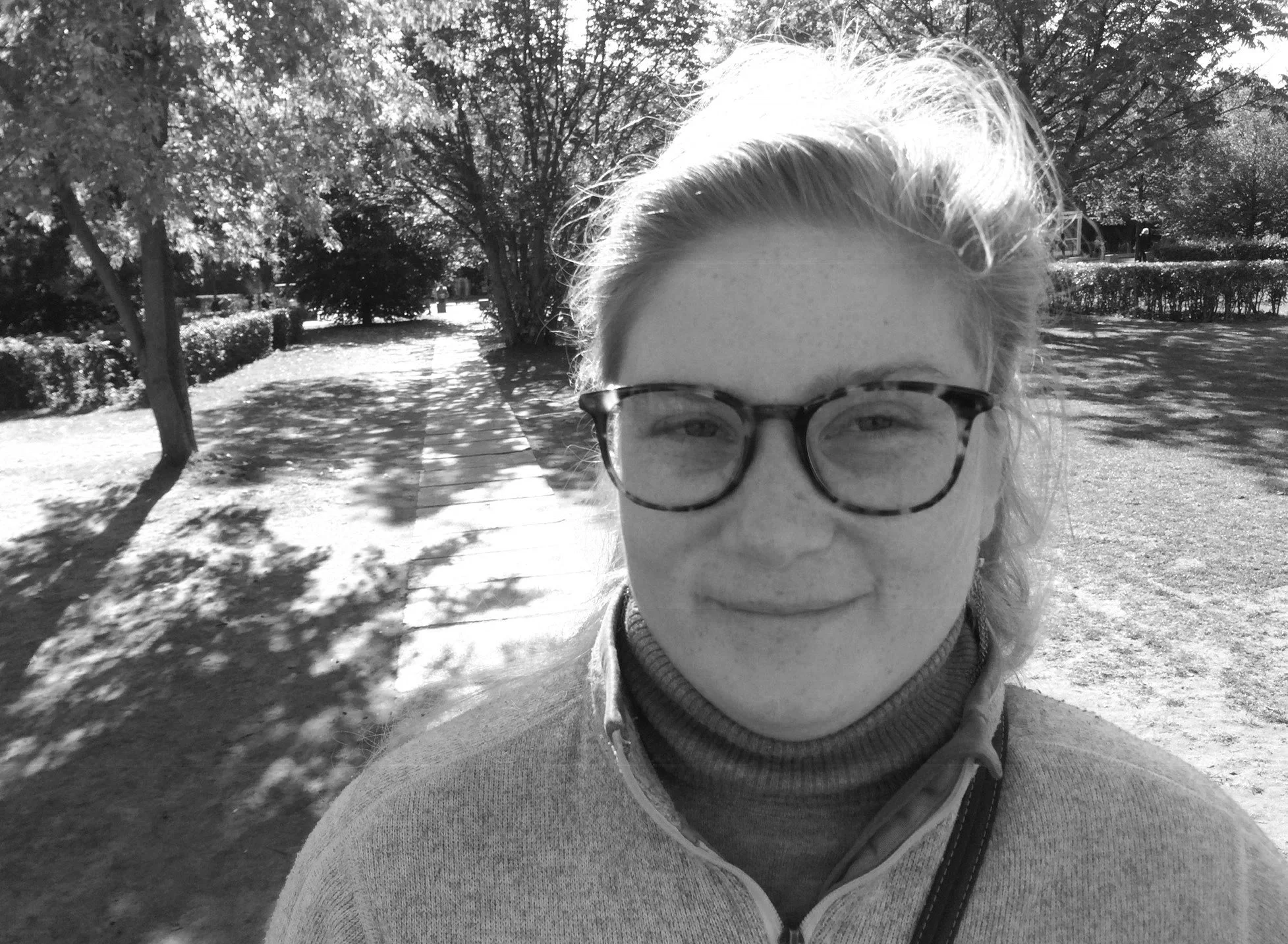 A black and white photo of a woman with short light hair, wearing glasses, standing outdoors in a park with trees, bushes, and a path visible in the background.