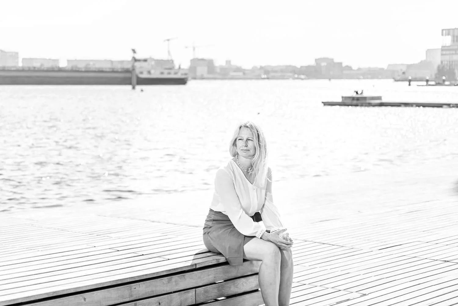 A woman sitting on a wooden dock by the water with ships and buildings in the background, black and white photo.