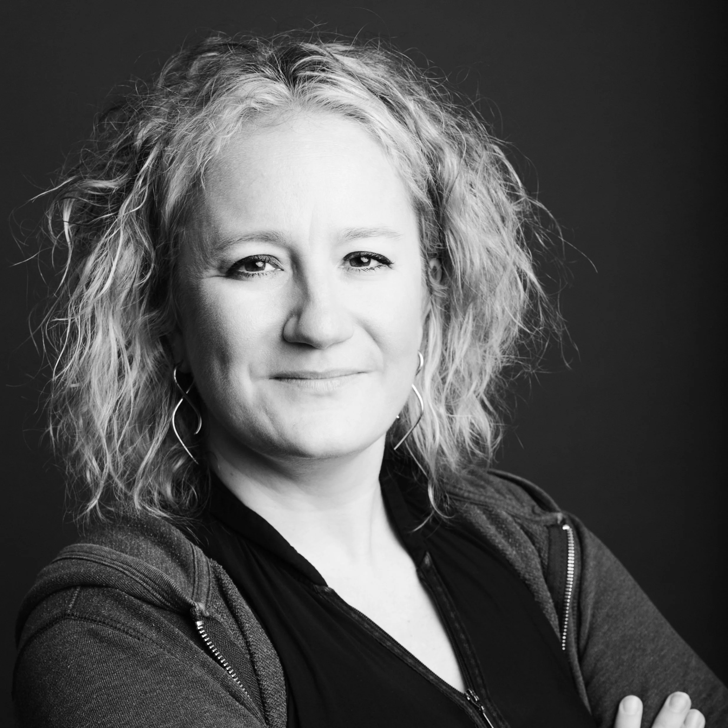 Close-up black and white photo of smiling woman with curly hair wearing a dark zip-up jacket, looking directly at the camera.