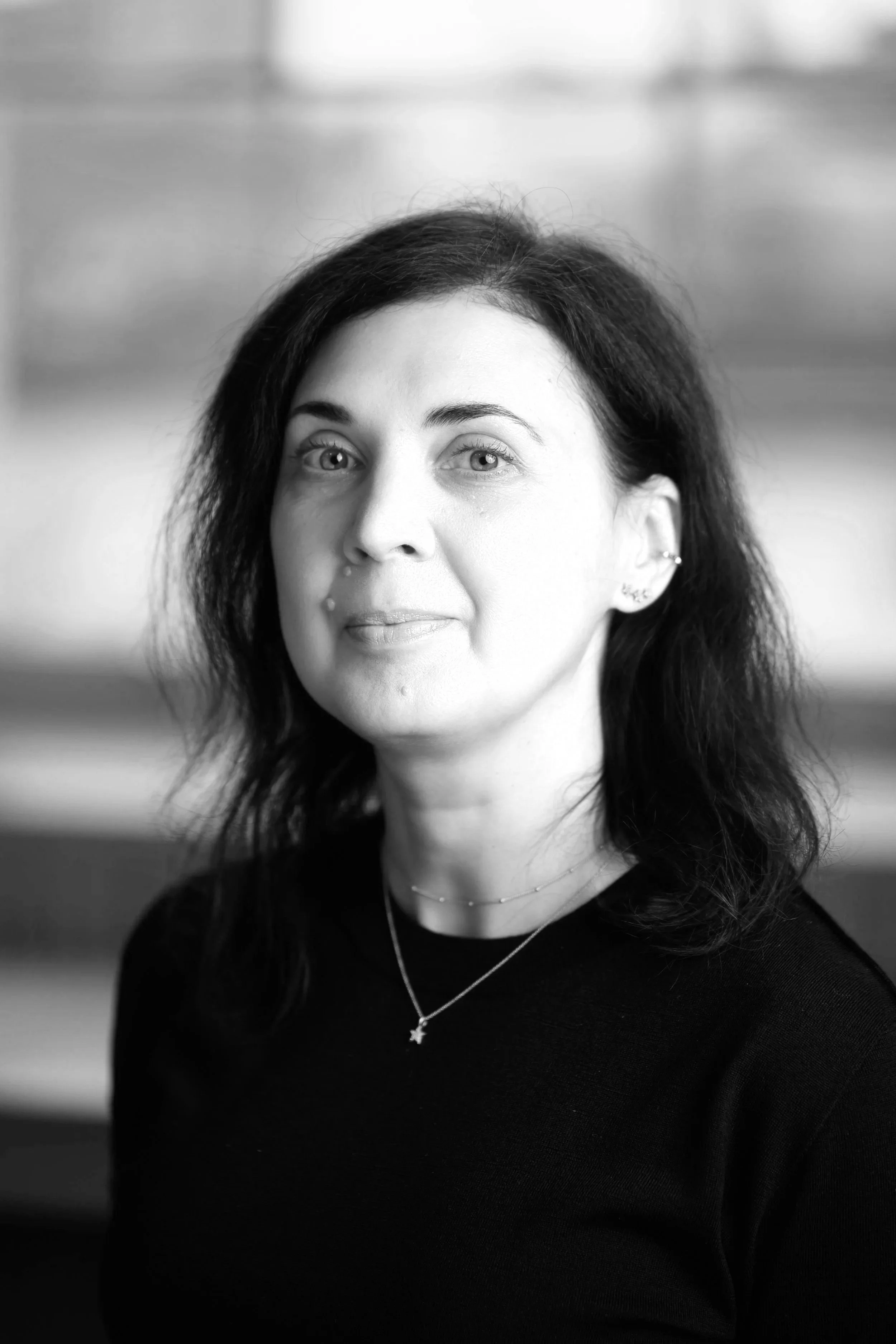 Black and white portrait of a woman with dark hair and light-colored eyes, wearing a necklace with a star pendant and a black top, looking slightly to the side.