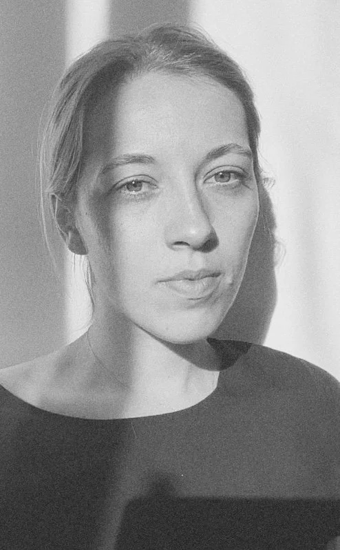 Black and white photo of a Nadja Schaetz with light hair, looking directly at the camera. She is wearing a dark top and is standing in front of a plain background with shadows cast on her face.