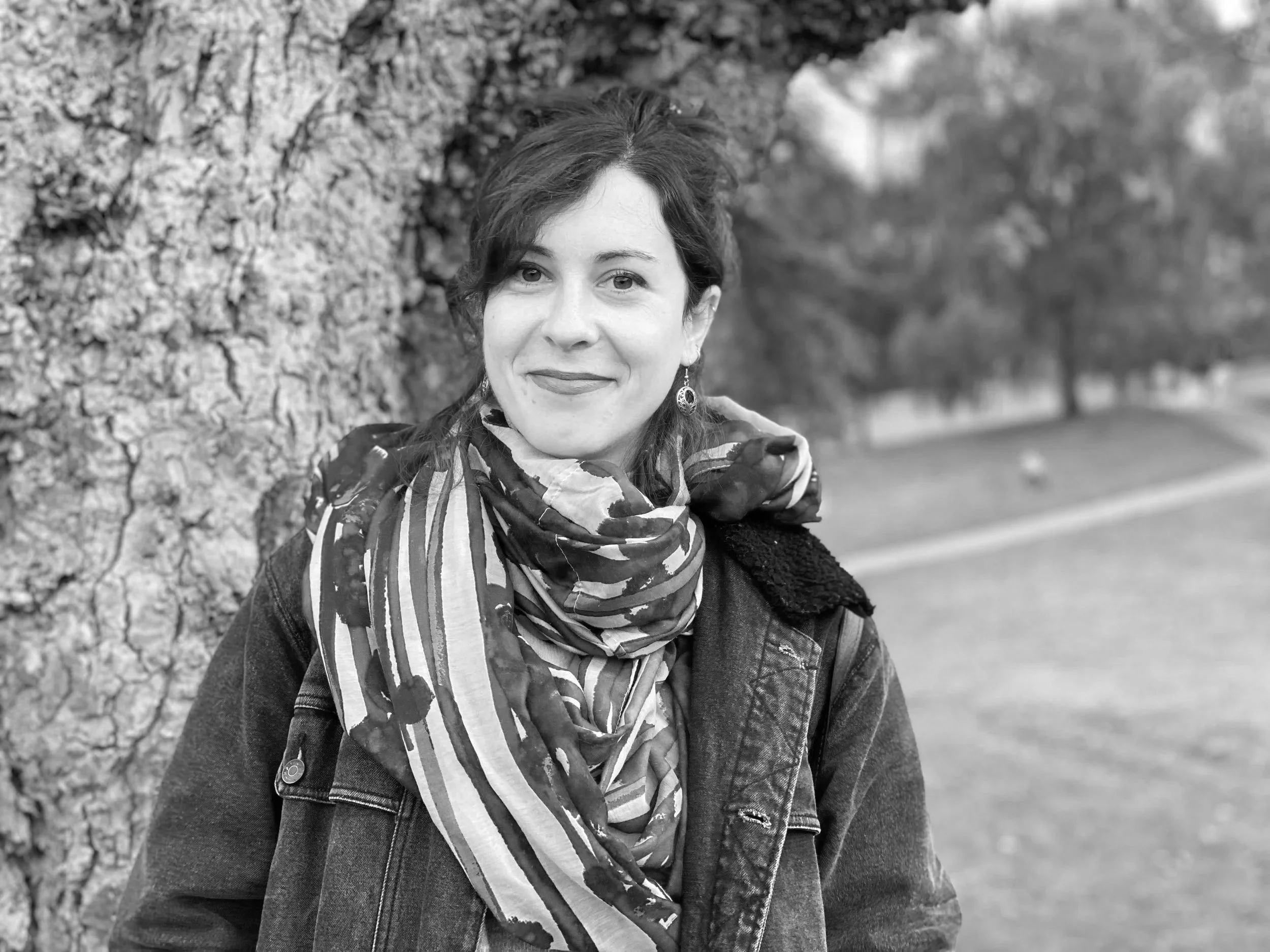 A woman with short, dark hair and earrings, smiling, leaning against a tree outdoors, wearing a scarf and a jacket, with blurred trees and grass in the background.
