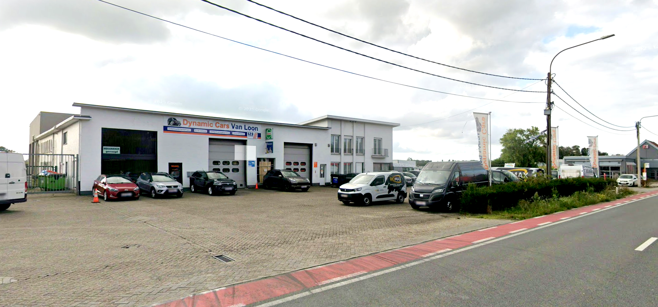 Een autogarage genaamd 'Dynamic Cars Van Loon' met verschillende auto's en bedrijfsvoertuigen op de parkeerplaats voor het gebouw, onder een bewolkte hemel.