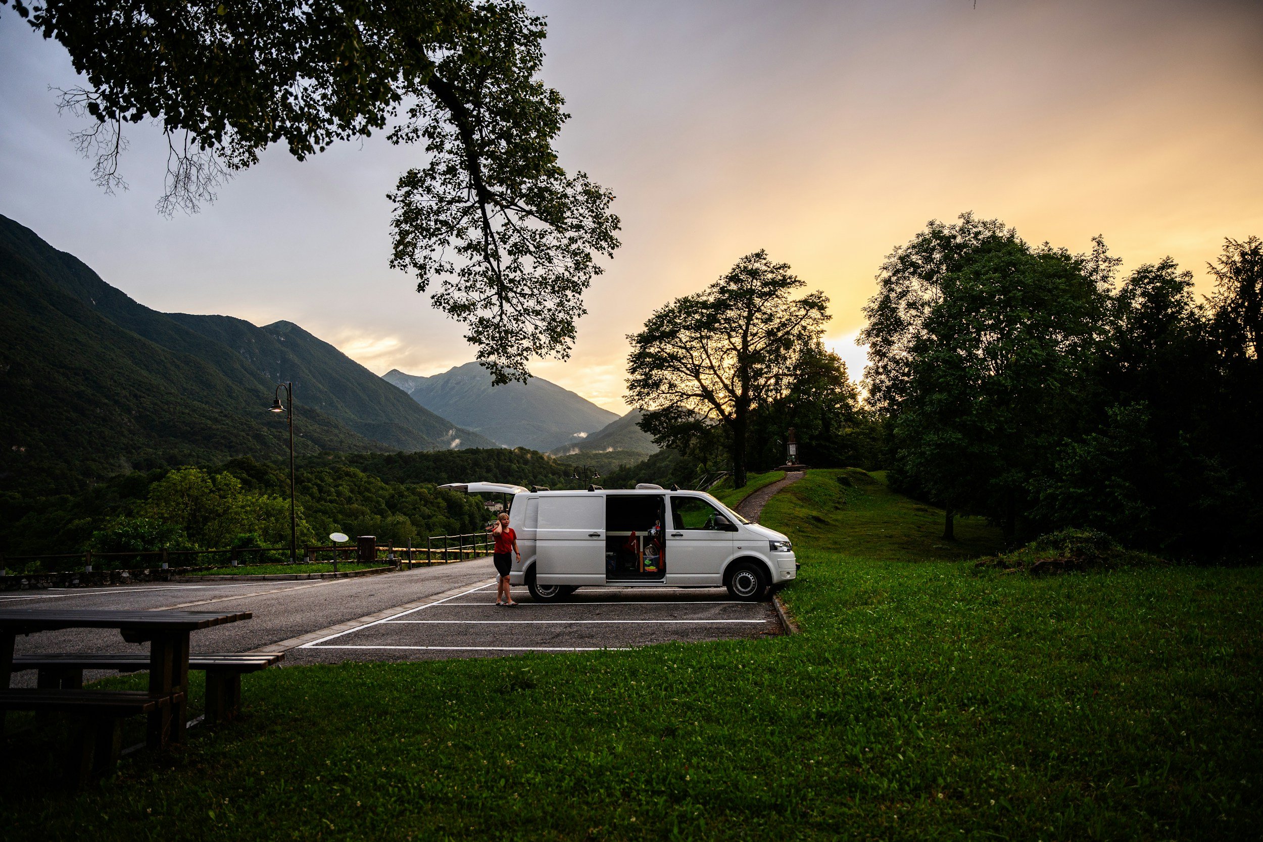 Een meisje in een rode jurk staat naast een witte bestelwagen in een bosrijke omgeving met bergen op de achtergrond, onder een zachtgekleurde hemel tijdens zonsondergang.