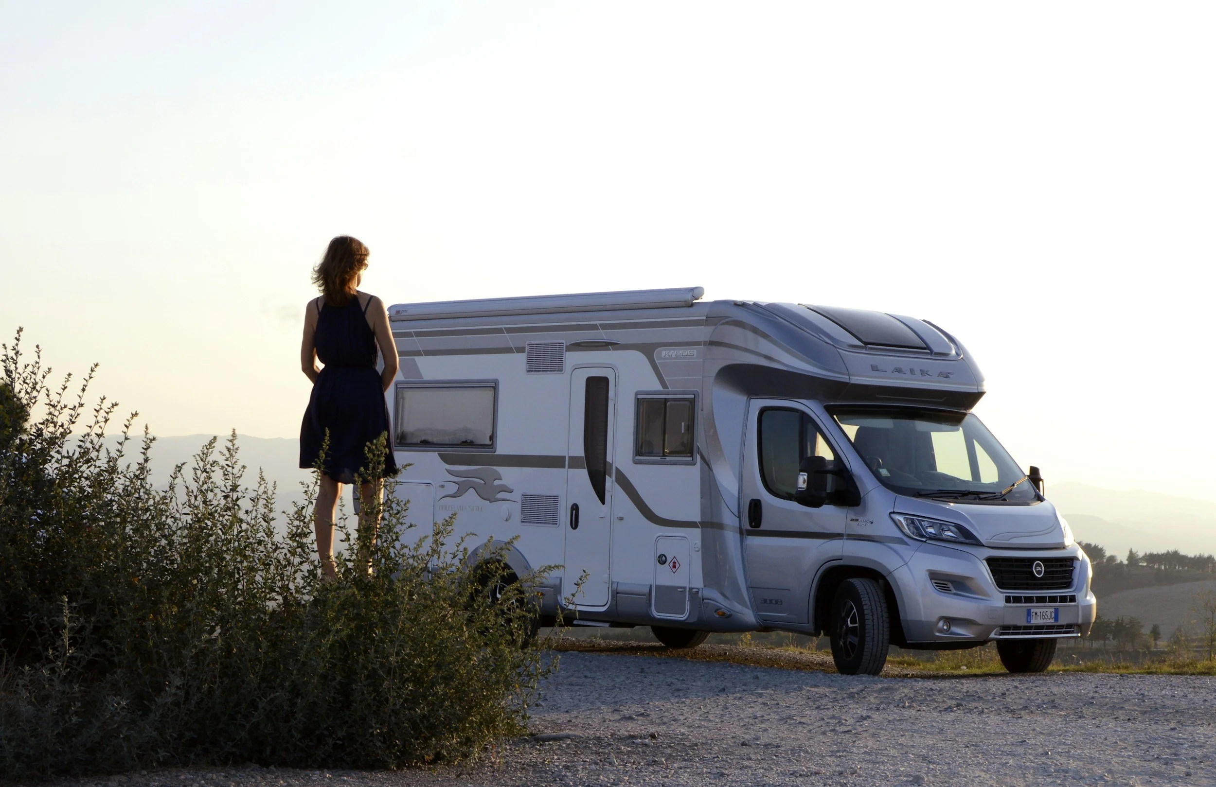 Vrouw staat naast een witte camper op een open veld bij zonsondergang, omgeven door struiken en met een bergketen op de achtergrond.