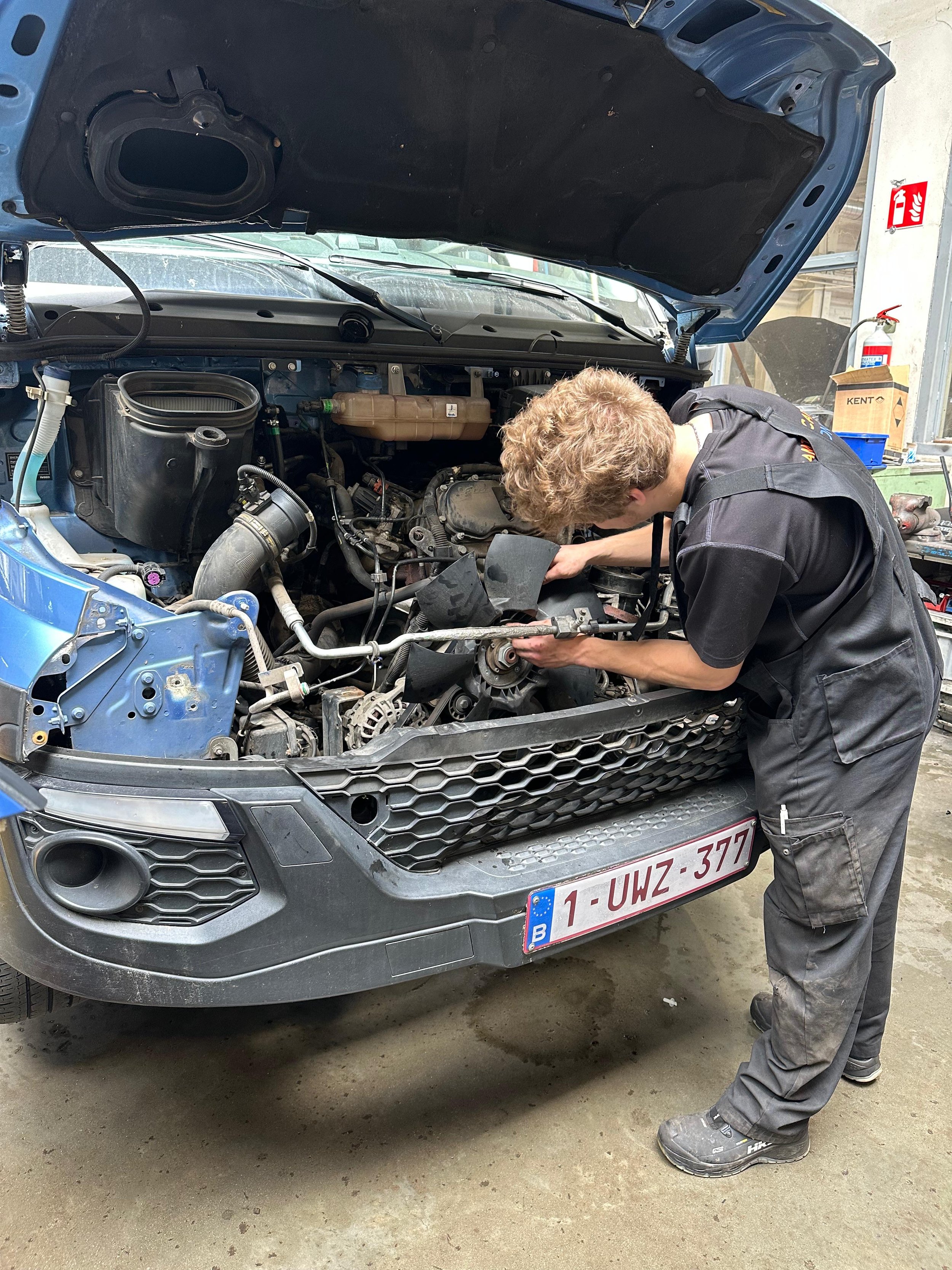 Een monteur werkt onder de motorkap van een grijze auto die in een garage staat. De motorkap is omhoog en de monteur is bezig met een reparatie of controle.