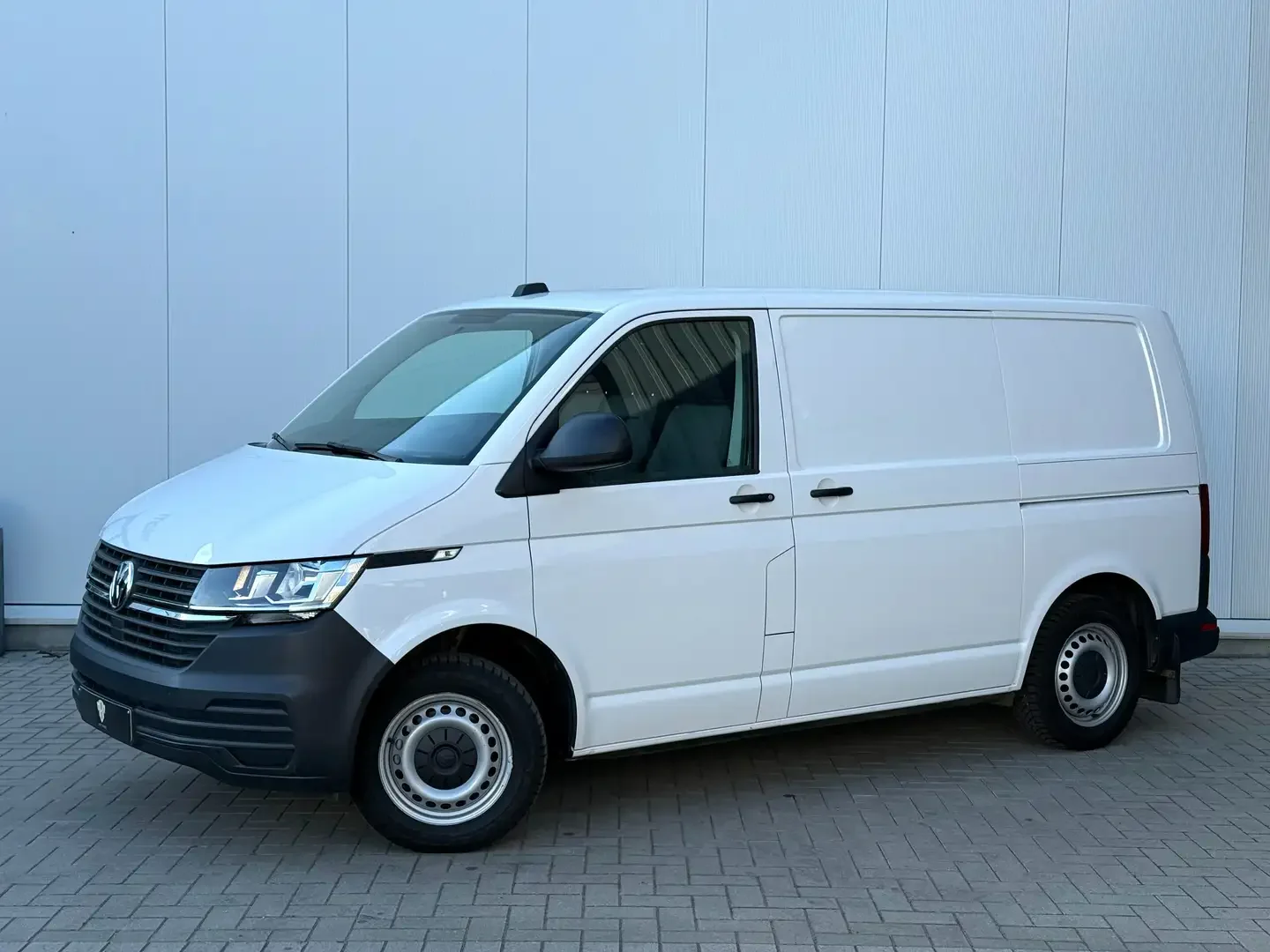 Witte Volkswagen bedrijfswagen geparkeerd op een betonnen oppervlak voor een lichtgrijze muur.