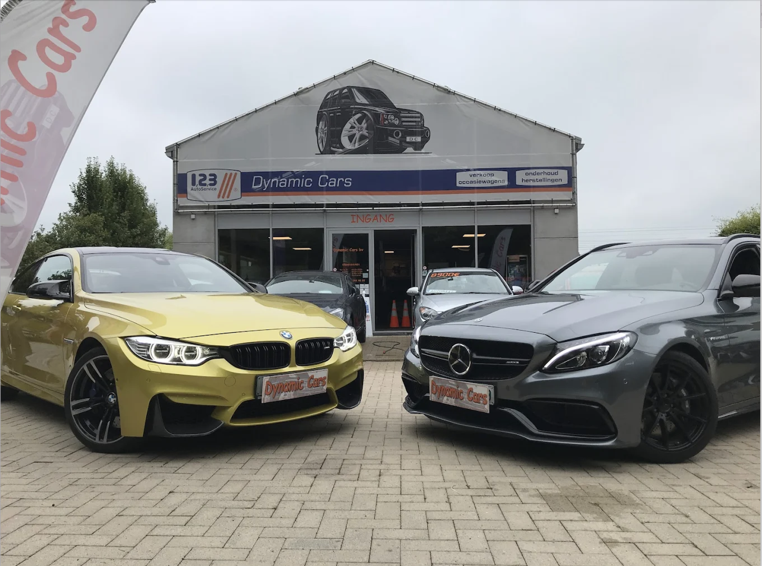 Een auto-expositie met verschillende sportauto's, waaronder een gele BMW en een zwarte Mercedes, voor een gebouw met een groot bord van een auto en reclameborden voor auto-onderhoud en verkoop.