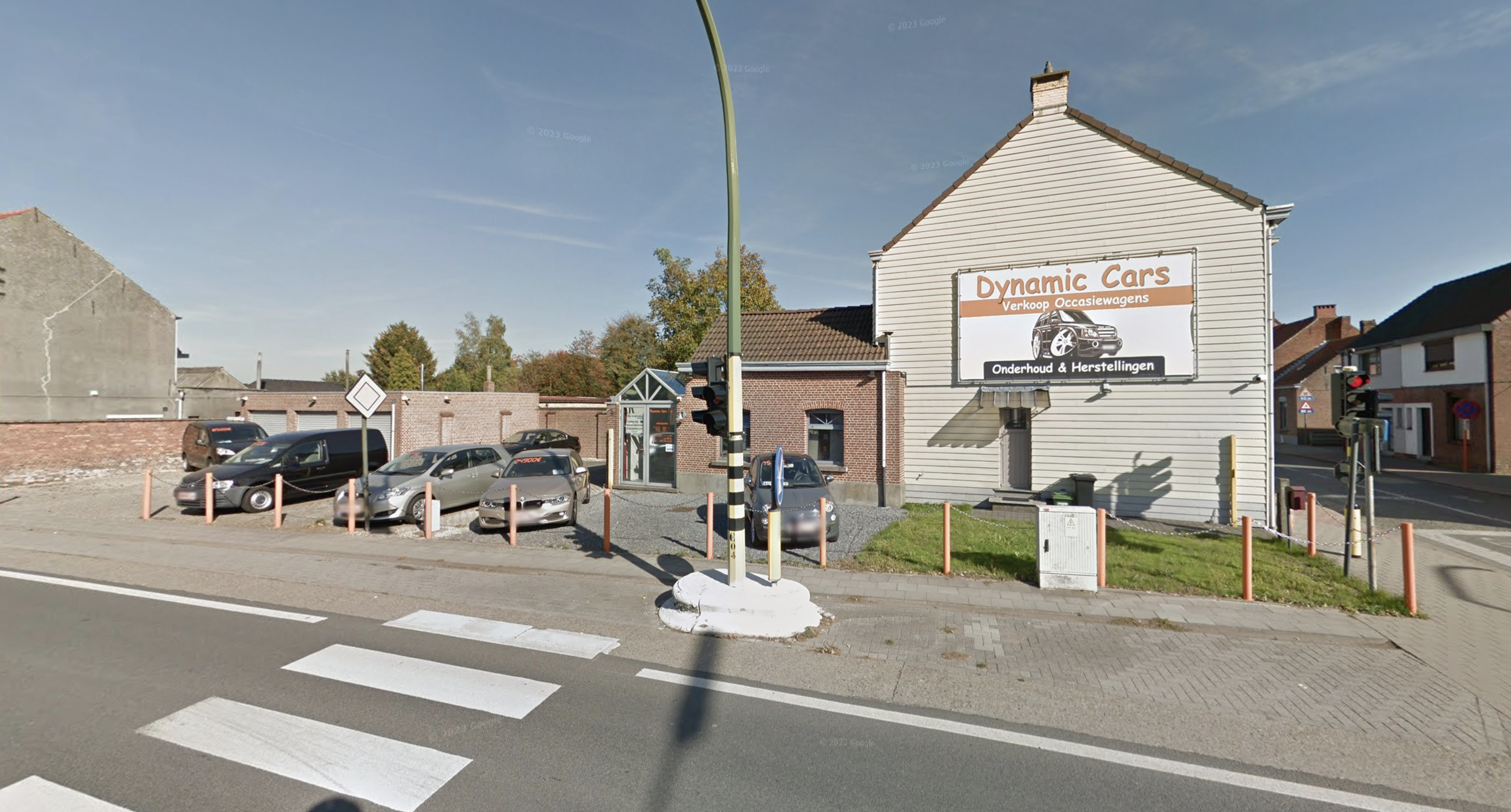 Winkelpand met reclamebord voor Dynamic Cars, autoservicebedrijf, met meerdere auto's geparkeerd voor het gebouw, verkeerslichten, zebra oversteekplaats en een parkeerplaats in een straat in een woonwijk.