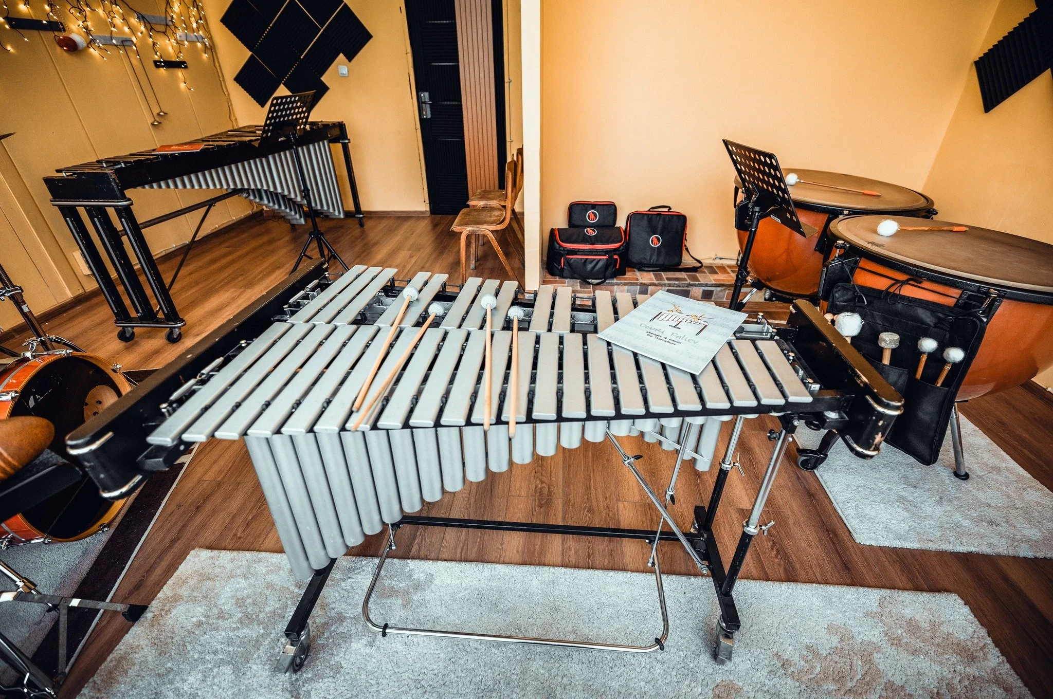 A musical practice room with a large xylophone in the foreground, a vibraphone to the left, and a pair of conga drums to the right. There are musical notes and a sheet of music on the xylophone. The room has beige walls, hardwood floors, and wall-mou