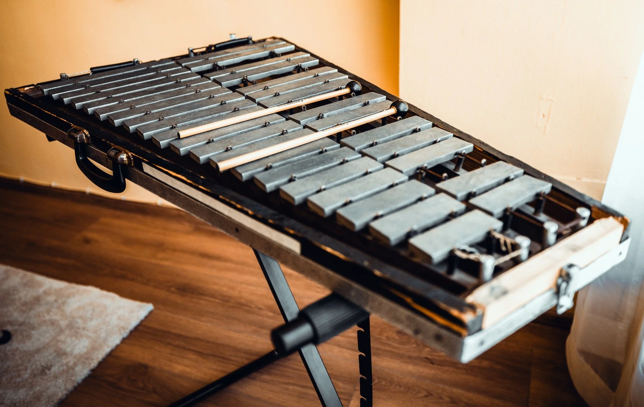 A xylophone with metal bars and mallets resting on top, placed on a stand in a room with hardwood flooring and a beige wall.