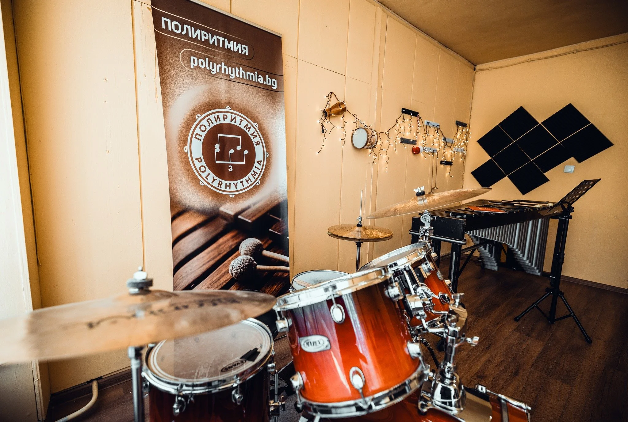 A room decorated for music, with a banner displaying a logo and text in Bulgarian, musical instruments like drums and a vibraphone, framed by string lights and soundproofing panels on the wall.