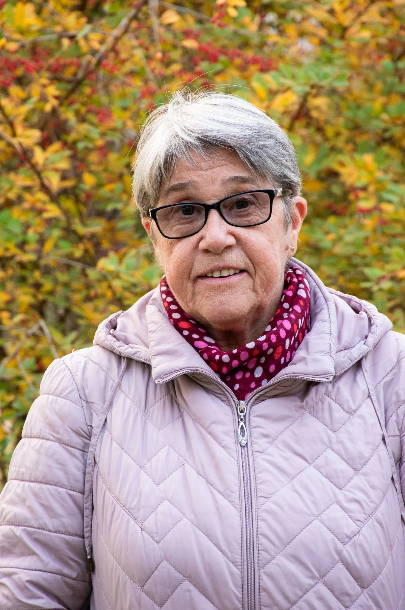 Close-up of an older woman with short gray hair, black glasses, wearing a pink quilted jacket and a red polka dot scarf, outdoors during autumn with colorful fall leaves in the background.