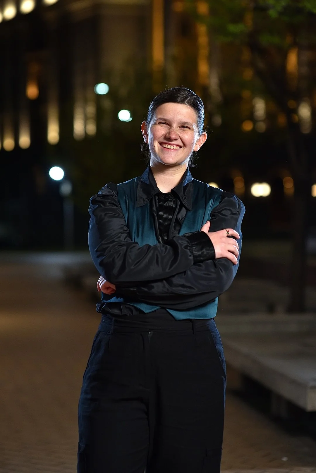 A young woman with dark hair in a ponytail, smiling with crossed arms, standing outdoors at night with blurred lights and trees in the background.