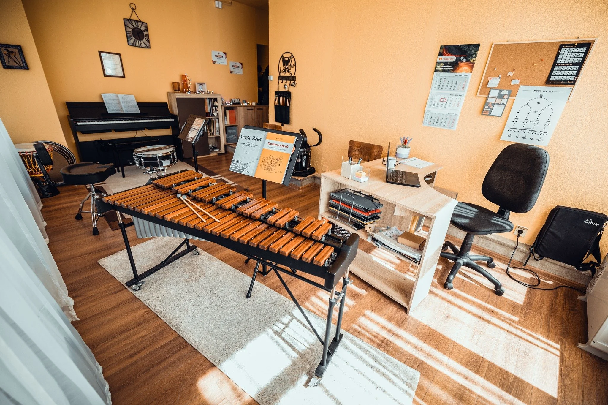 A music practice room with a wooden xylophone, a piano, a music stand, and a desk with a laptop on it. The room has orange walls with framed pictures, a bulletin board with notes, and a calendar. There is also a chair, a small bookcase, and a drum se