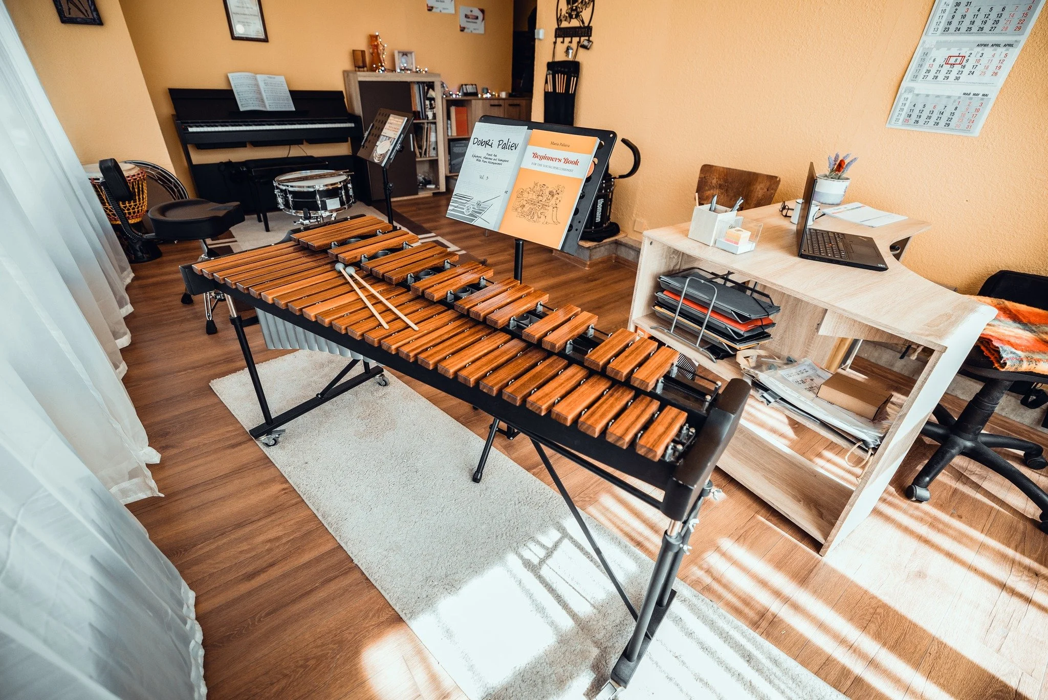 A room with a wooden floor and yellow walls, featuring a xylophone in the foreground, a music stand with sheet music, a piano with open sheet music, and a desk with a laptop, pen holder, and various papers. There are calendars on the wall and a books