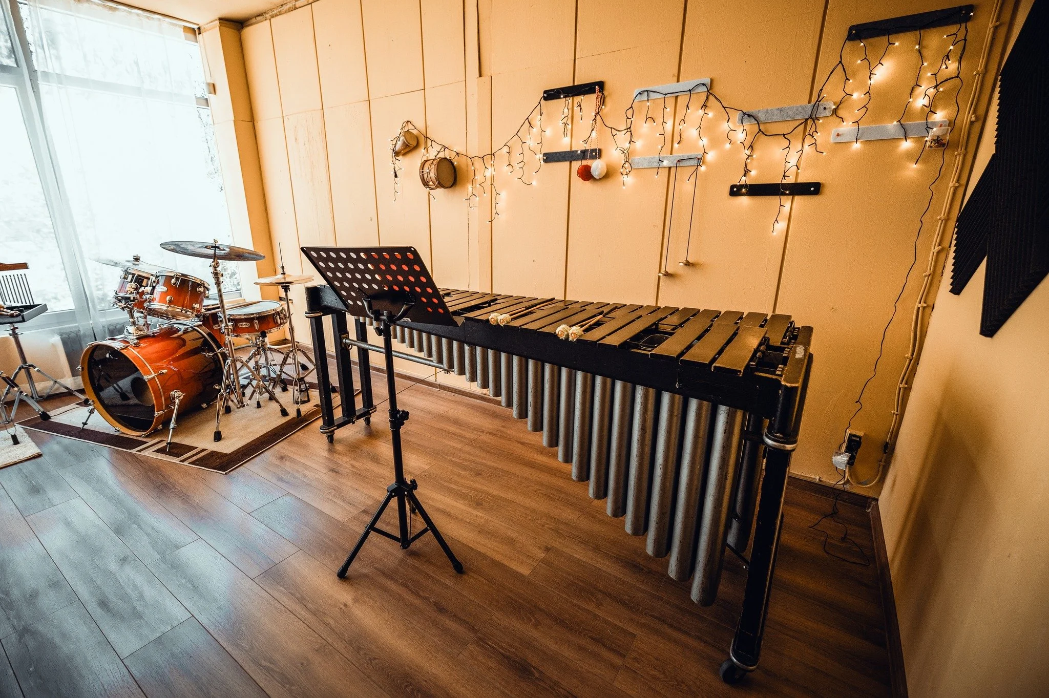 A music rehearsal room with a drum set, a xylophone, a music stand, and string lights on the wall. There is a window on the left side of the room letting in natural light.