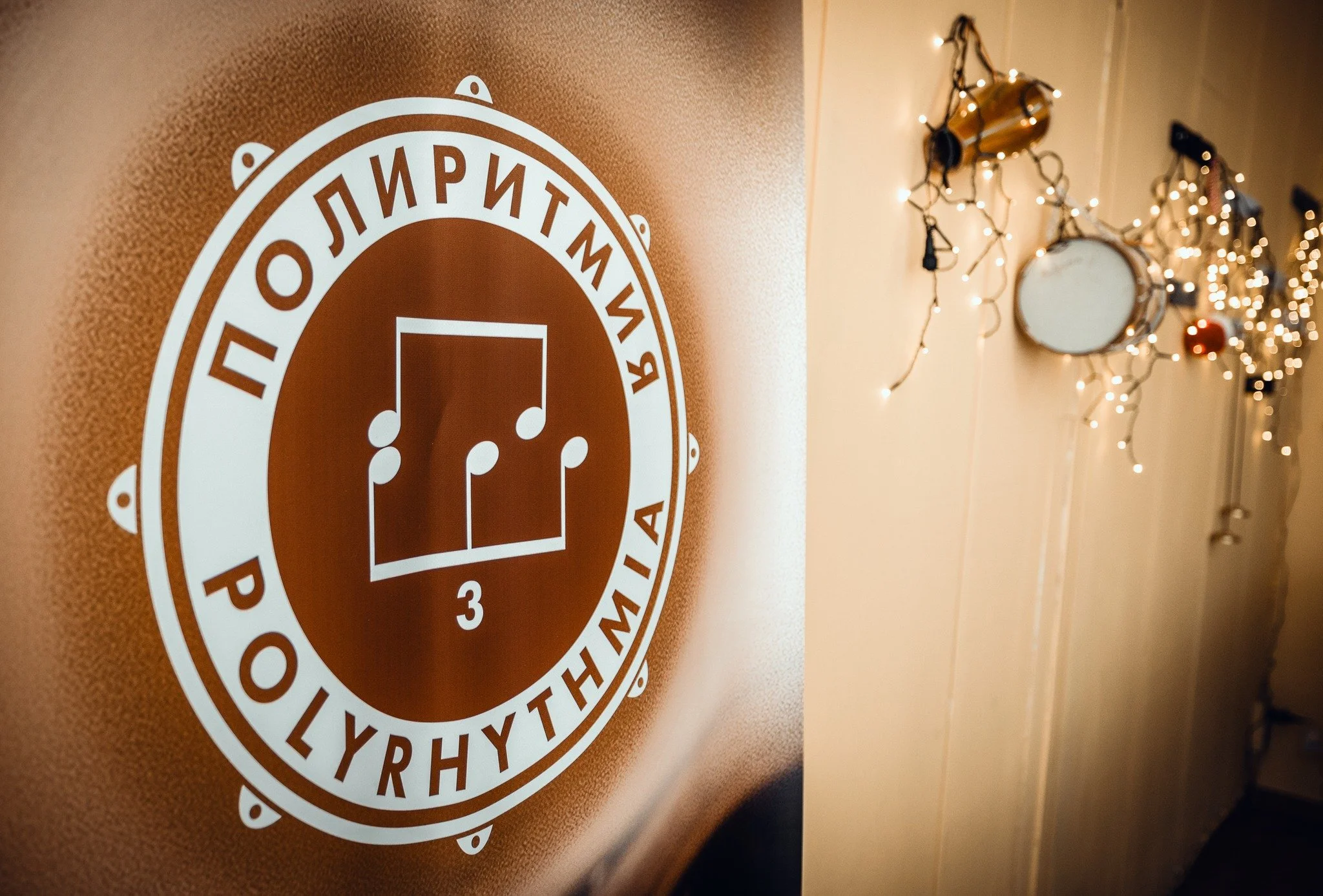 A close-up of a sign with white text and symbols on a brown background, slightly out of focus, and decorated walls with holiday lights in the background.