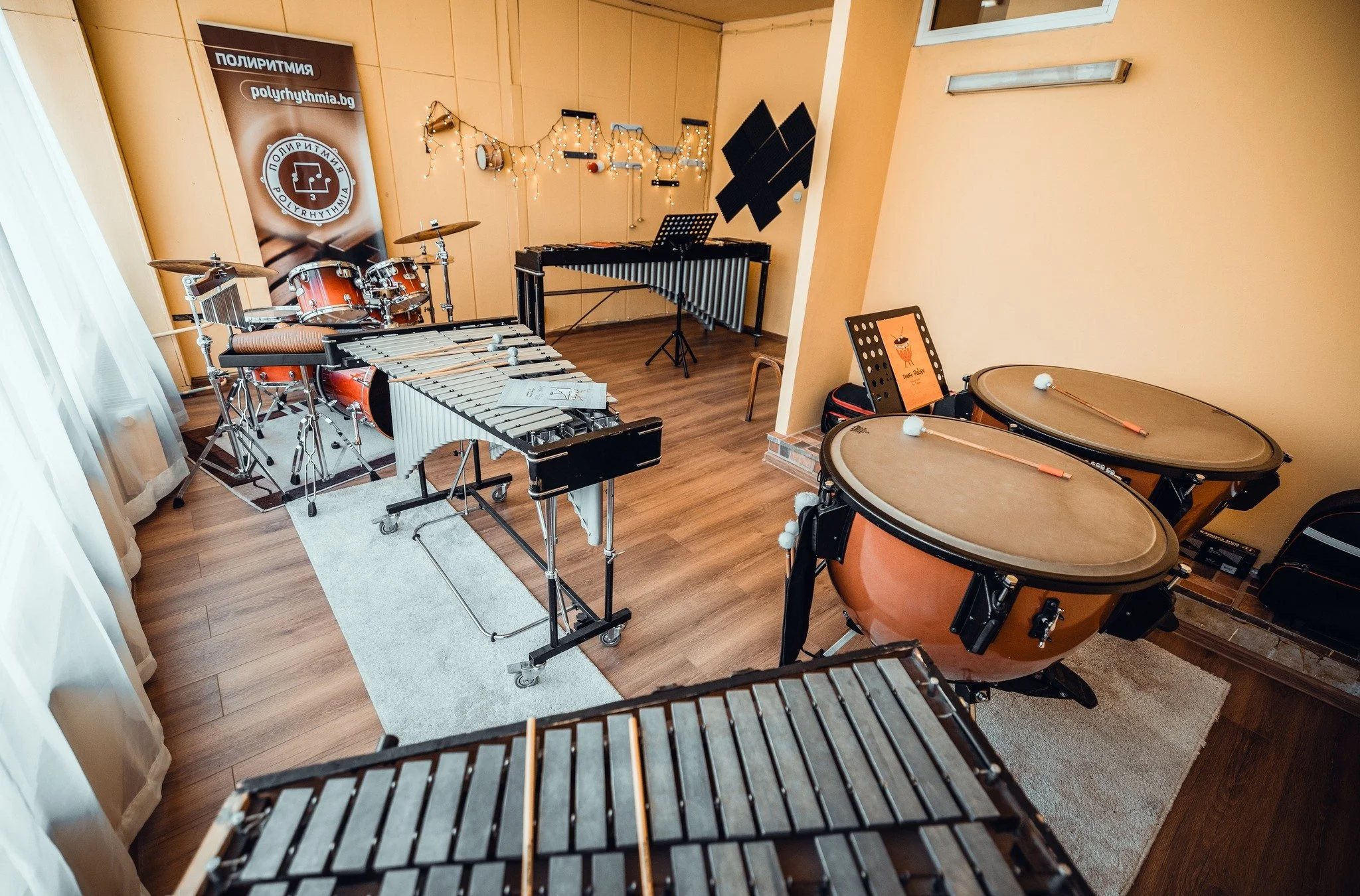 Music practice room with drums, xylophones, percussion instruments, music stands, and black soundproofing panels, decorated with string lights.