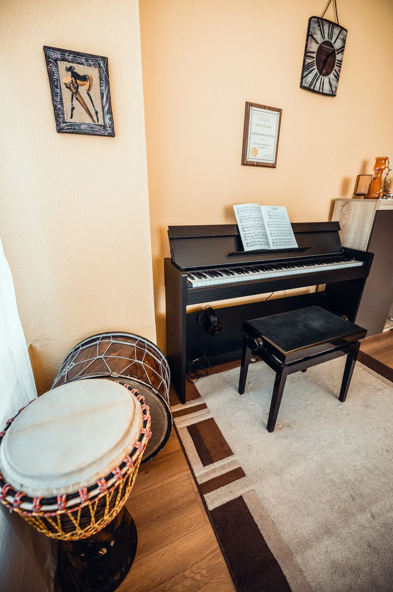 A black digital piano with sheet music on its music stand, positioned against a beige wall with framed artwork and a clock. There is a partially visible drum in the foreground and a decorative wind chime or sculpture on the right.
