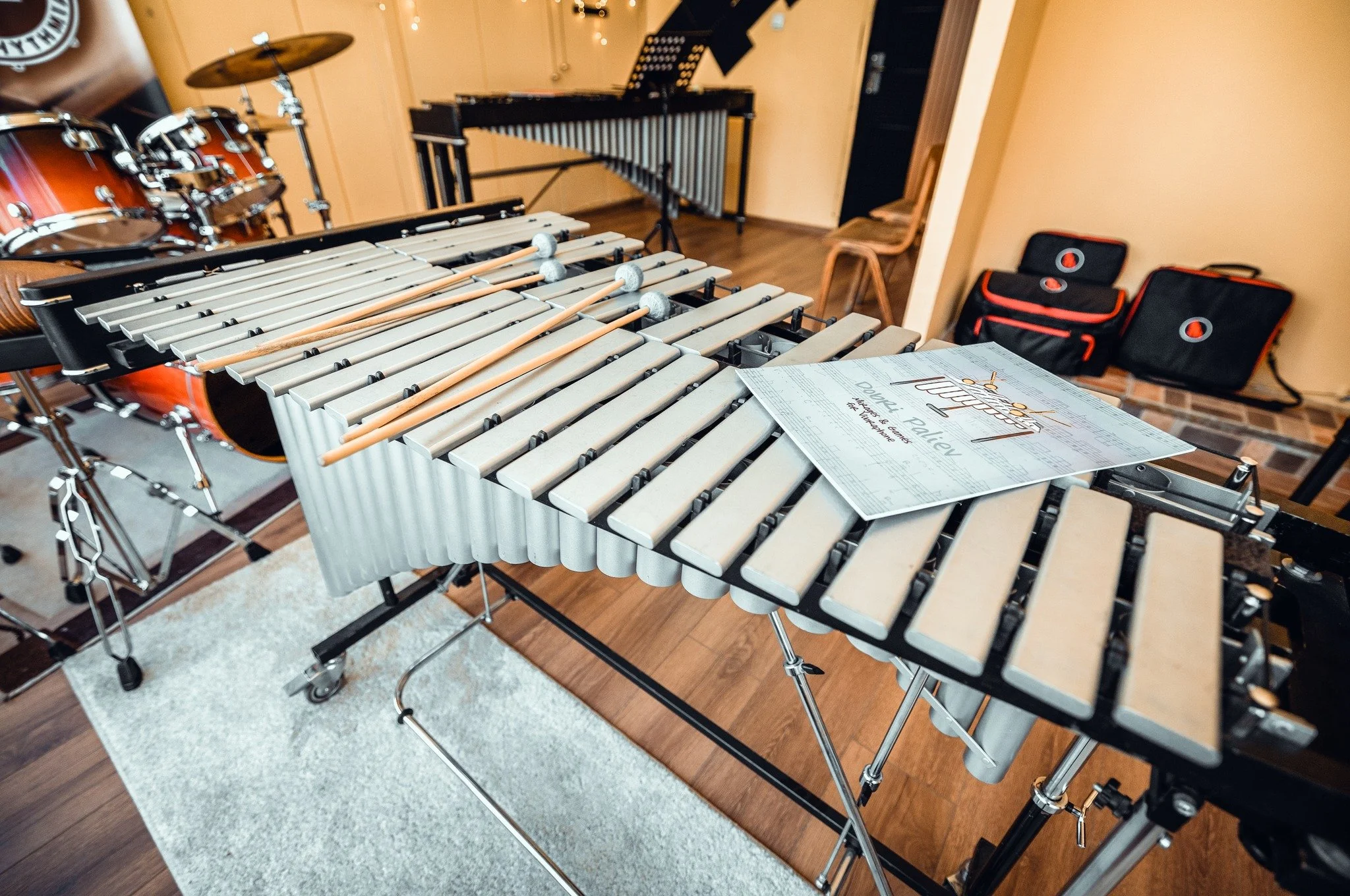 A room with musical instruments including a xylophone with mallets on top, a drum set, and a vibraphone. There are chairs and music bags in the background.
