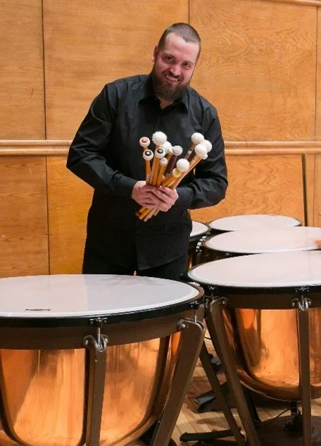 A man holding drumsticks with mallets standing among large percussion drums in a music room.