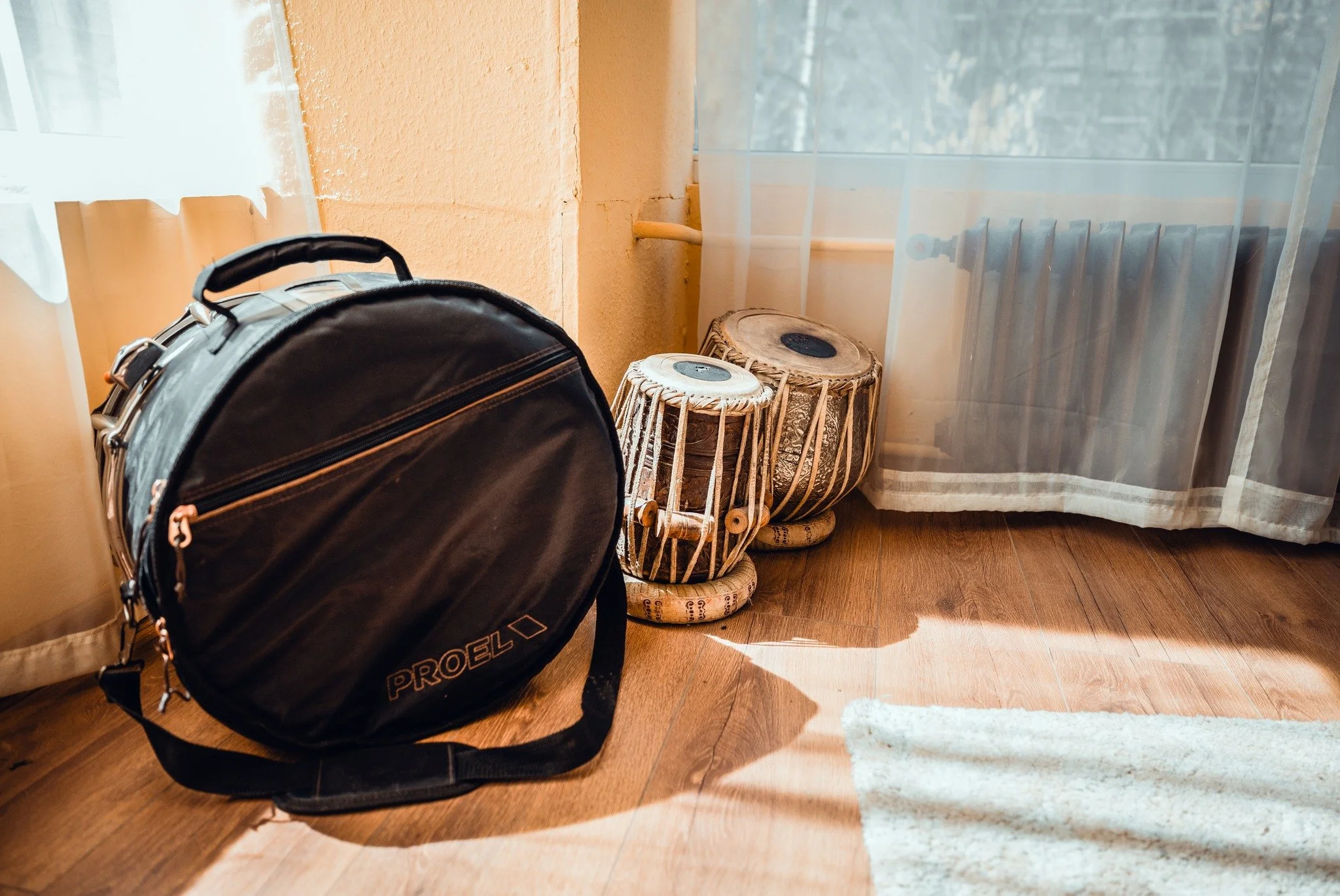 A black Proel backpack resting on a wooden floor next to two traditional drums, with a window with sheer curtains and a radiator in the background.