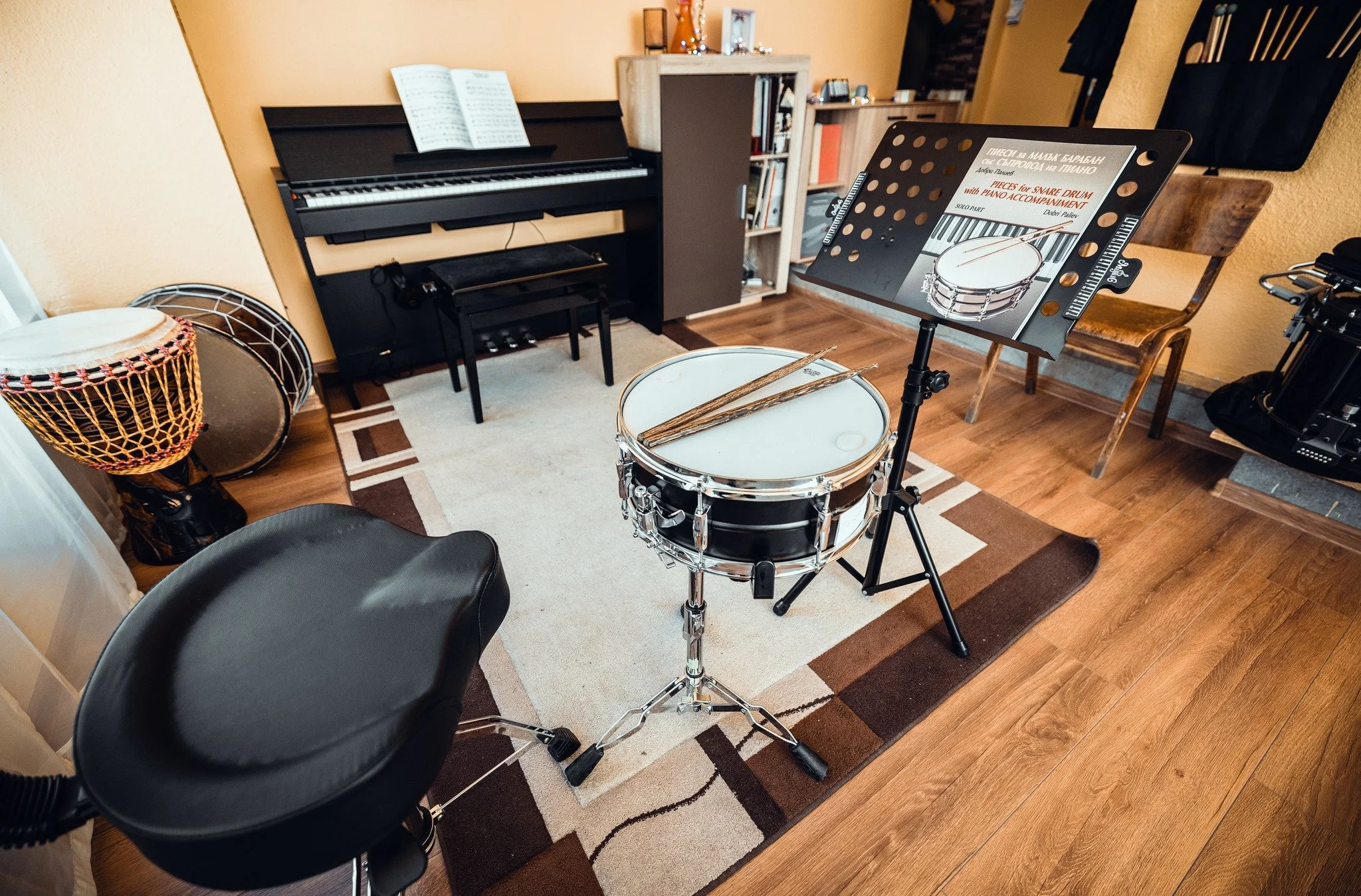 A music practice room with a digital piano, a music stand with sheet music, a snare drum with drumsticks, a drum stool, a large drum, a black chair, a small rug, and some bookshelves holding books and decorative items, with hardwood flooring and yell