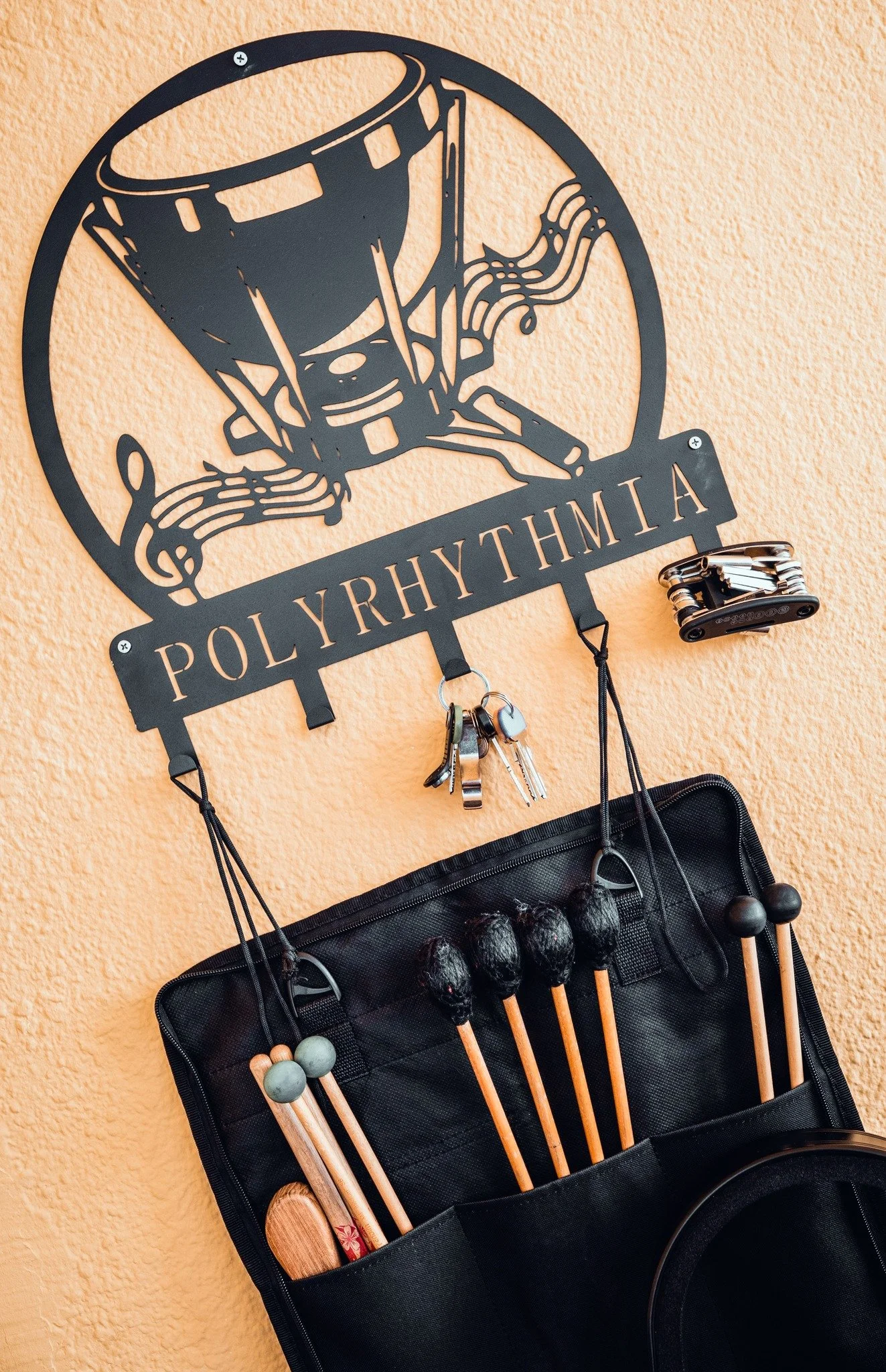 A metal wall decor with the word POLYRHythMIA and a drum design, hanging on an orange wall, with a keychain holder and a wooden eyebrows brush hanging underneath.