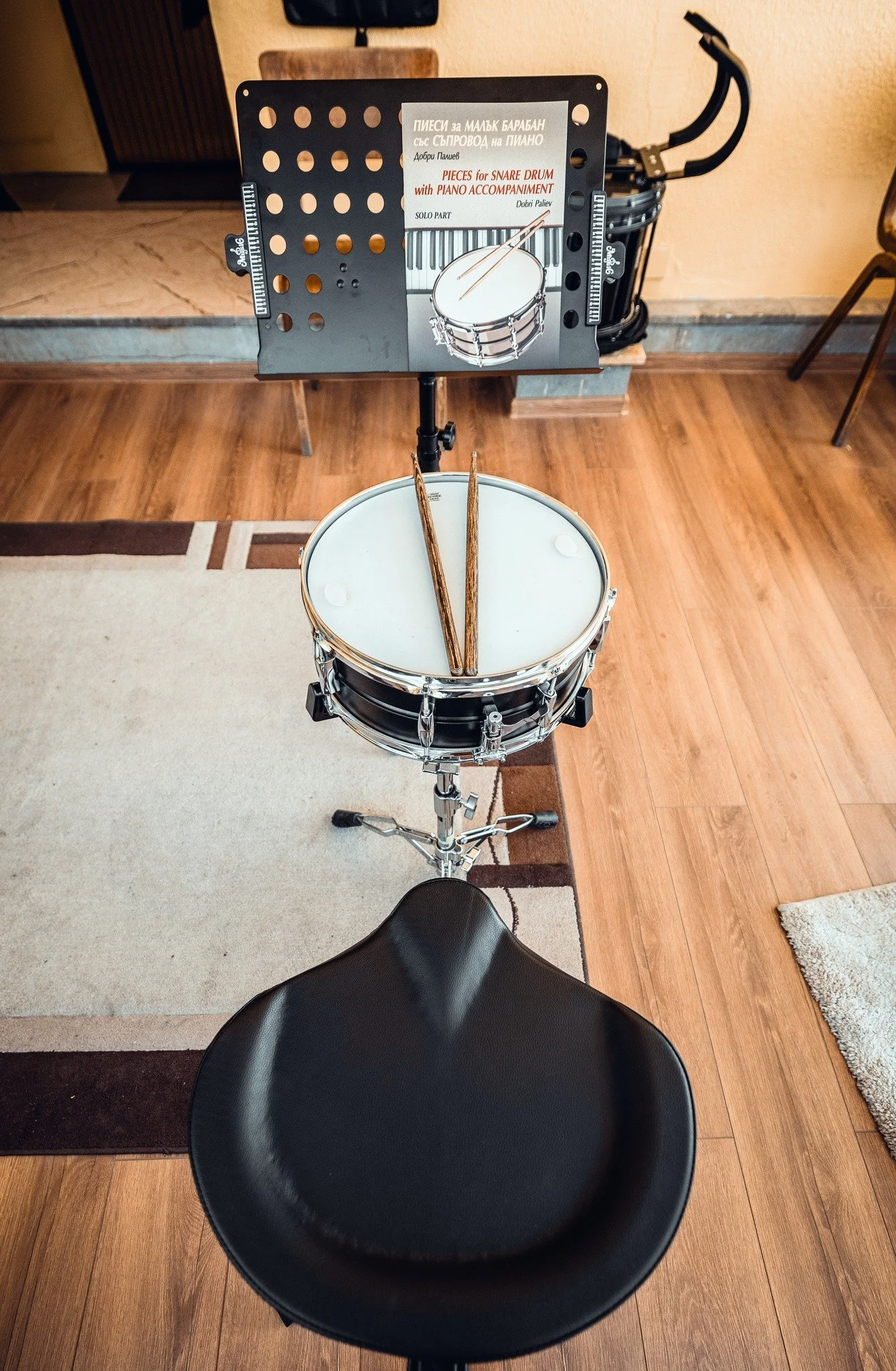 A music stand with a sheet of paper that depicts a snare drum and piano keys, with a pair of drumsticks on it, positioned in front of a black drum and a black chair in a room with wood flooring.