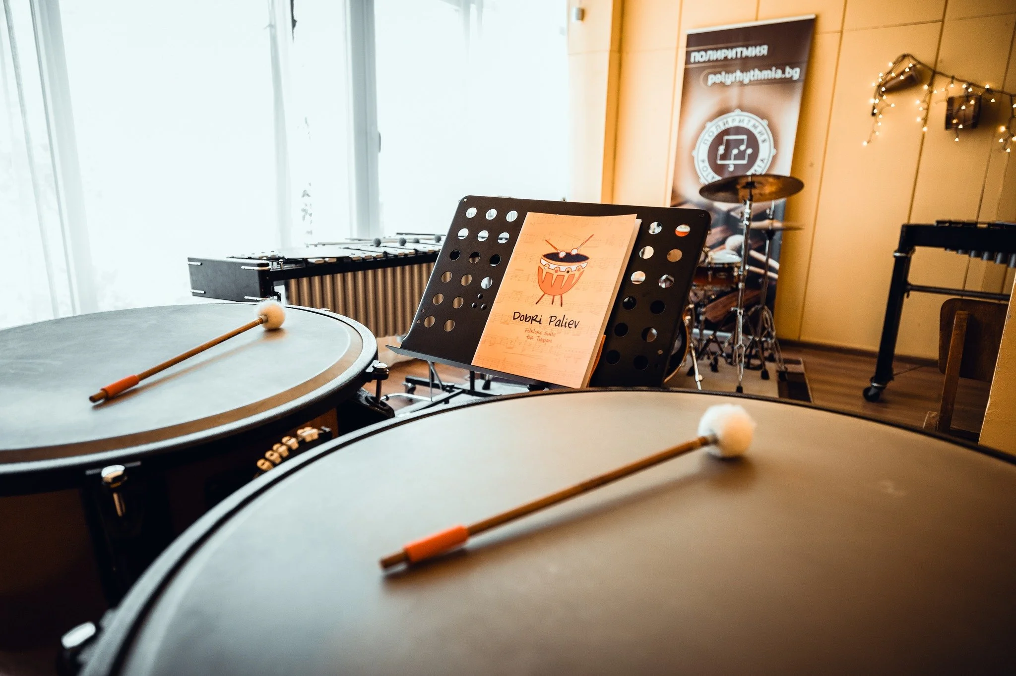 Music stand with a sheet of music and a sign that reads "Dobri Paliev" in a room with musical instruments, including a xylophone, drums, and a partial view of a drumstick in the foreground.