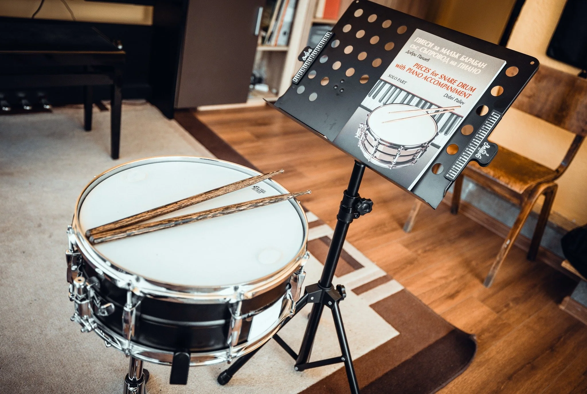 A music stand with sheet music titled 'Pieces for Snare Drum with Piano Accompaniment' positioned beside a snare drum with drumsticks on top, set in a room with wooden flooring and a bookshelf in the background.