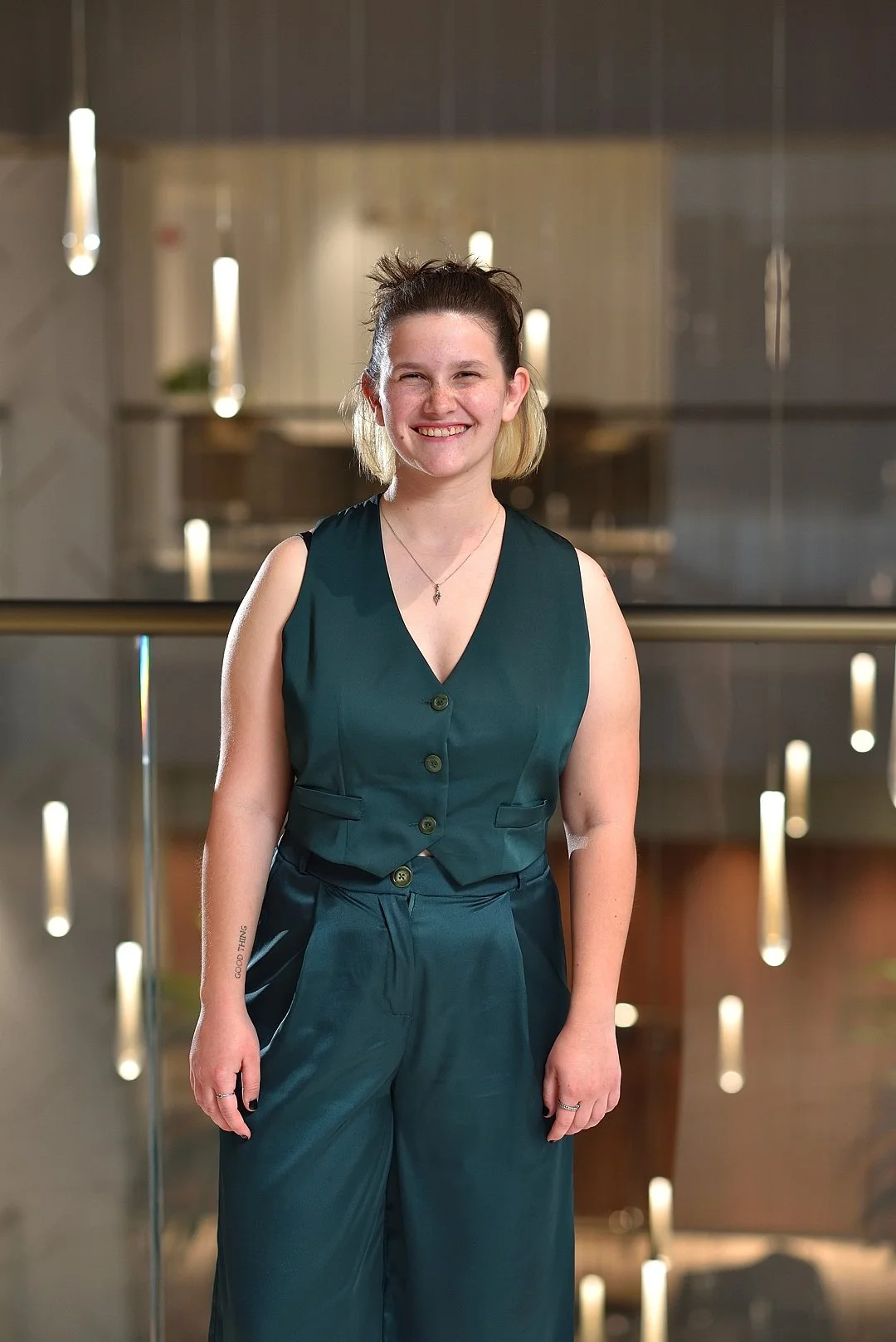 A young woman with short hair, smiling, standing in front of a glass railing with lights behind her. She is wearing a dark green sleeveless vest and matching wide-leg pants.