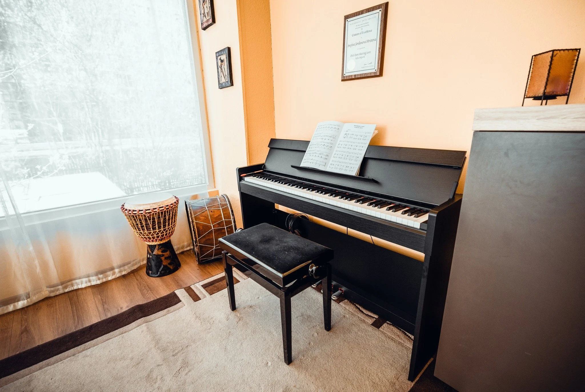 A black digital piano with sheet music on its stand, a small black bench in front, facing a window with sheer curtains, and framed artwork and a certificate hanging on a peach-colored wall. Two drums, a tall drum with a woven body and a smaller drum,