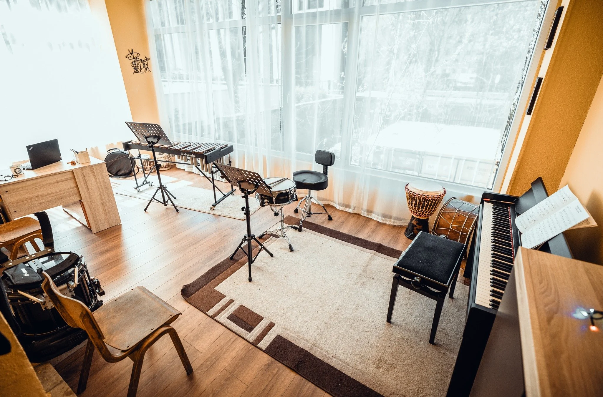 A cozy music room with large windows covered by light curtains, wooden floors, a black electronic keyboard, a music stand, a stool, a drum, and chairs, with sunlight illuminating the space.