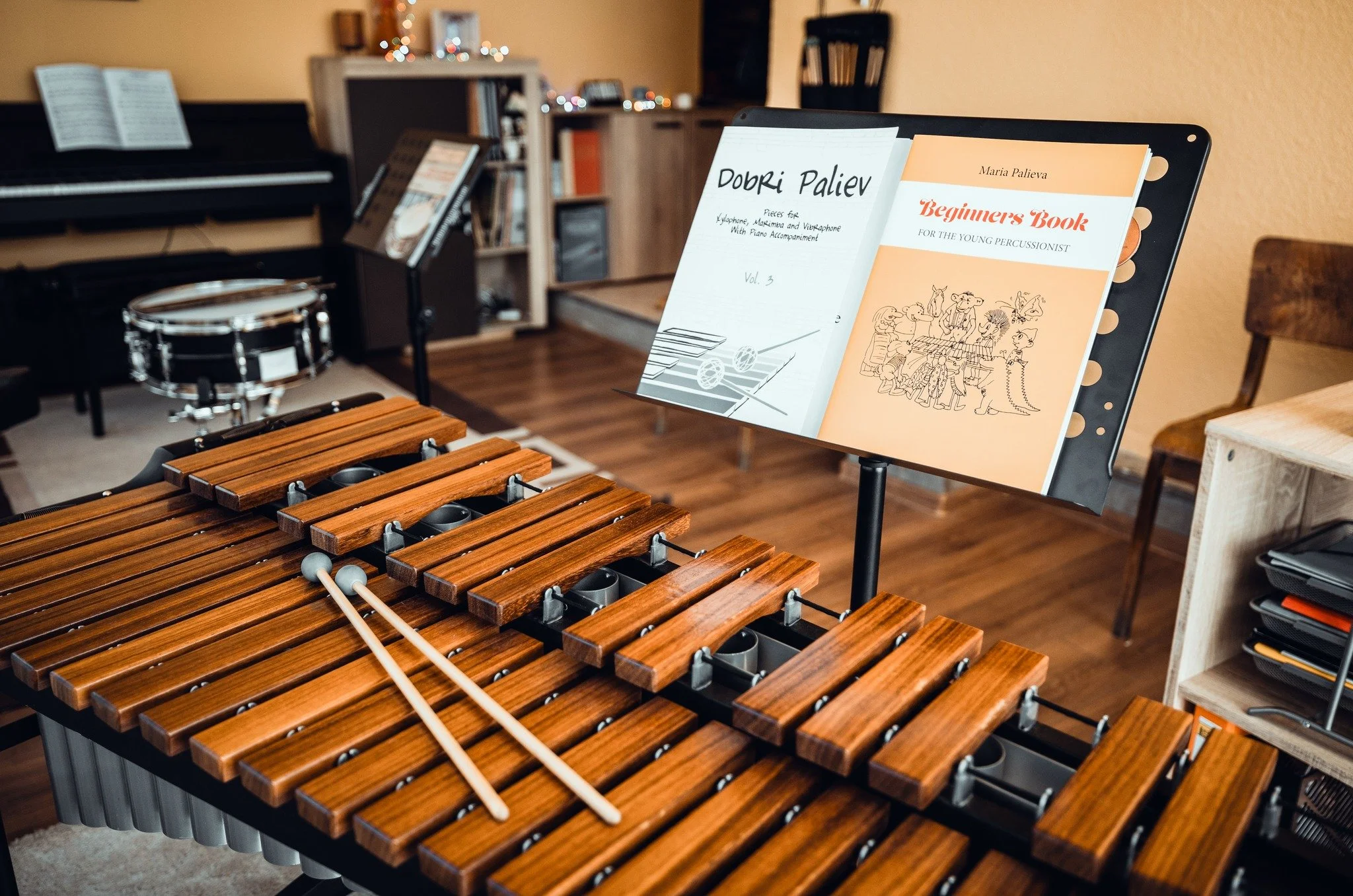 A music room with a wooden xylophone in the foreground, a music stand with sheet music titled 'Beginner's Book for the Young Percussionist', a piano, a snare drum, and various bookshelves with books in the background.