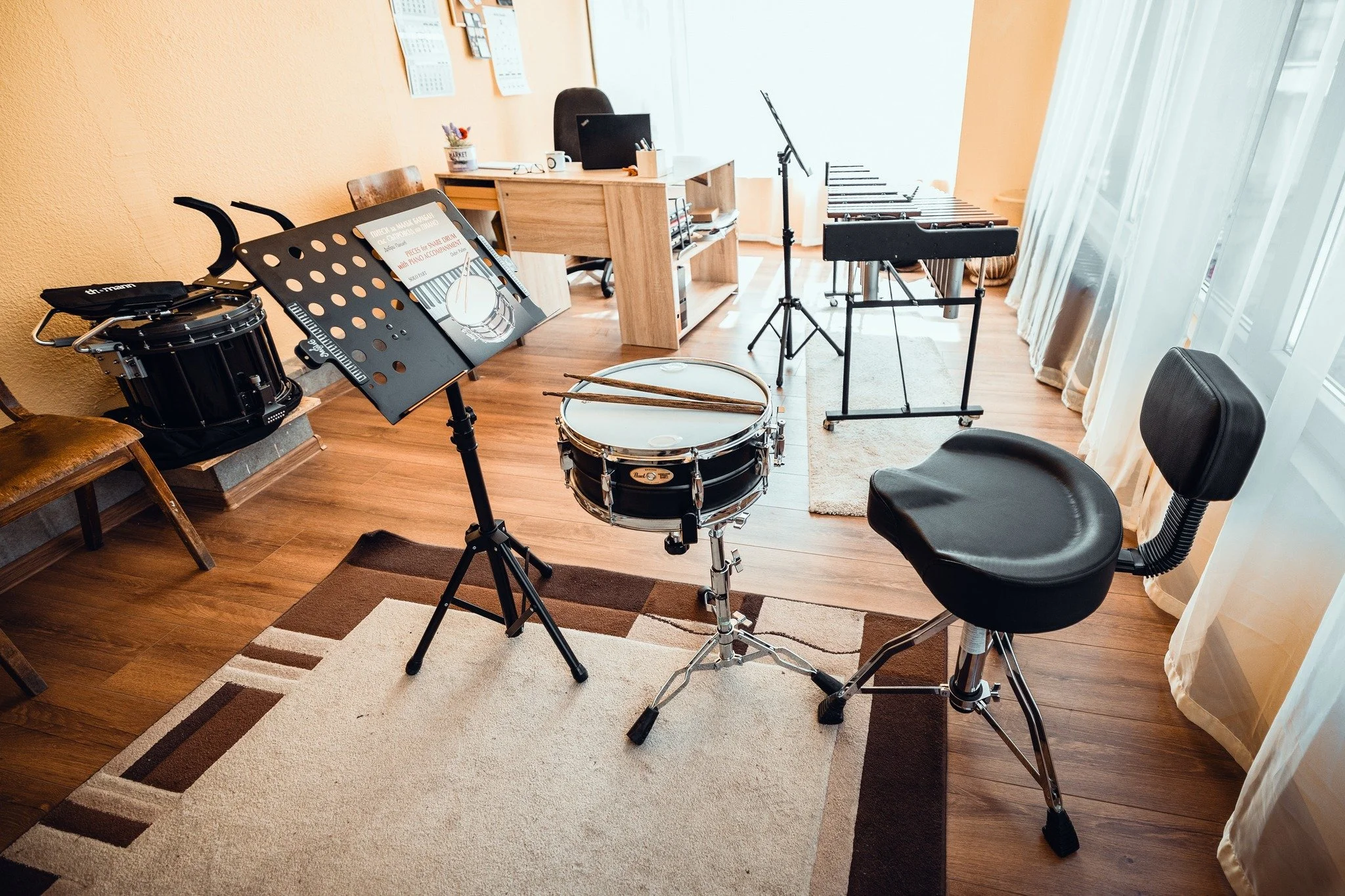 A music room with percussion instruments including a drum, a music stand with sheet music, a chair, a xylophone, a music stand, a computer desk, and a bookshelf, near a large window with curtains.