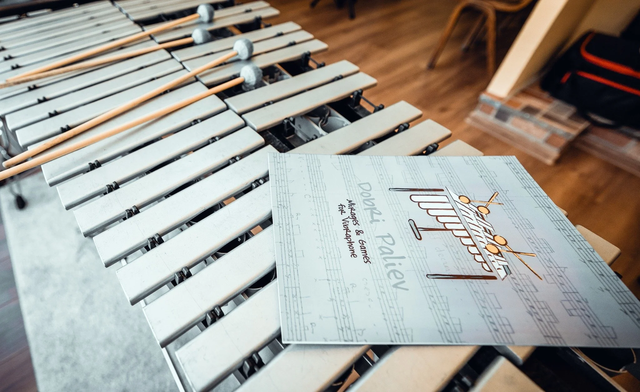 A xylophone with several mallets resting on top of its bars, and a leaflet titled 'Party Planning' with handwritten notes and musical staff background, on a table in a room with hardwood flooring and a chair.